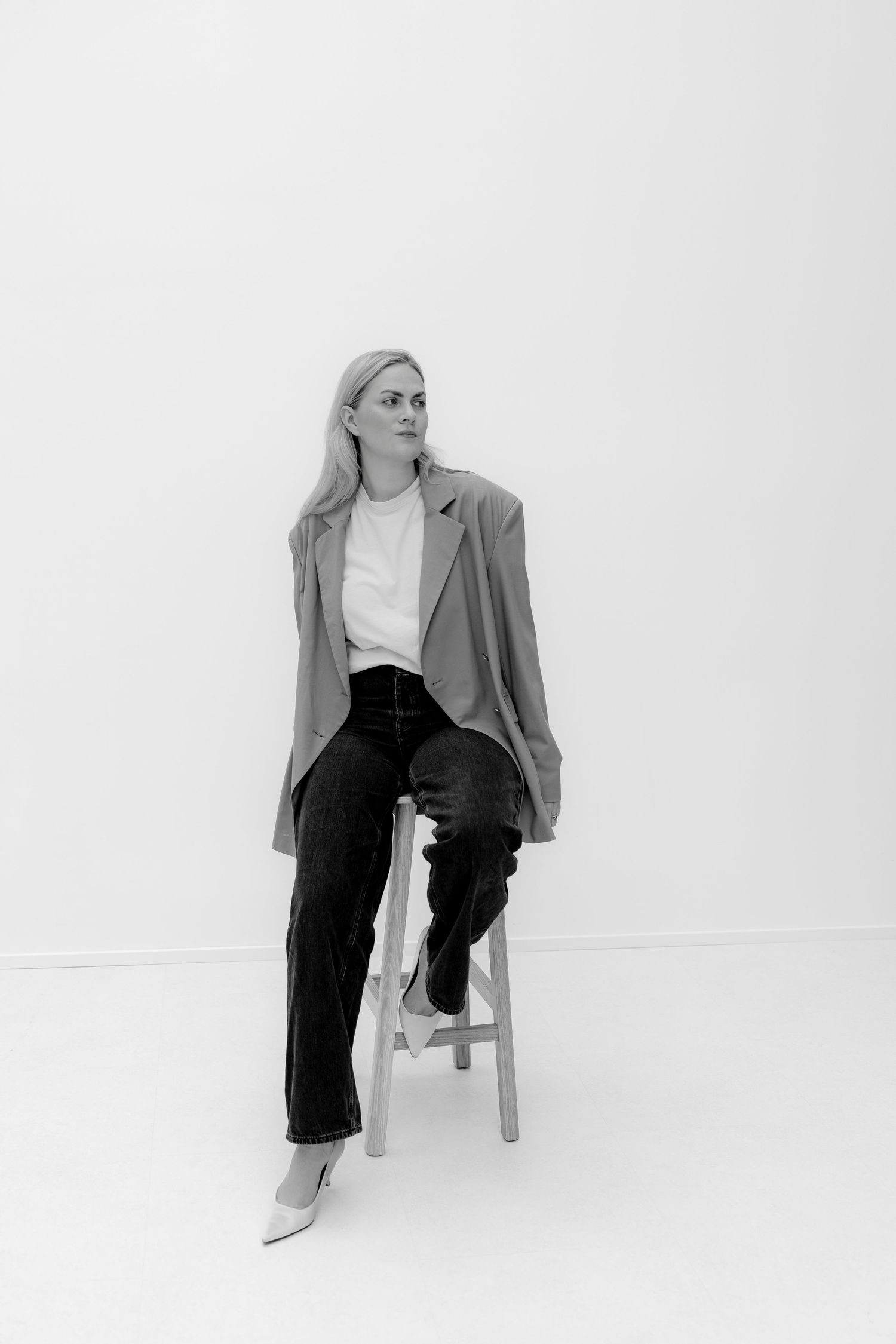 Black and white photo of a blonde woman in blazer and jeans sitting on a wooden stool against a white background.