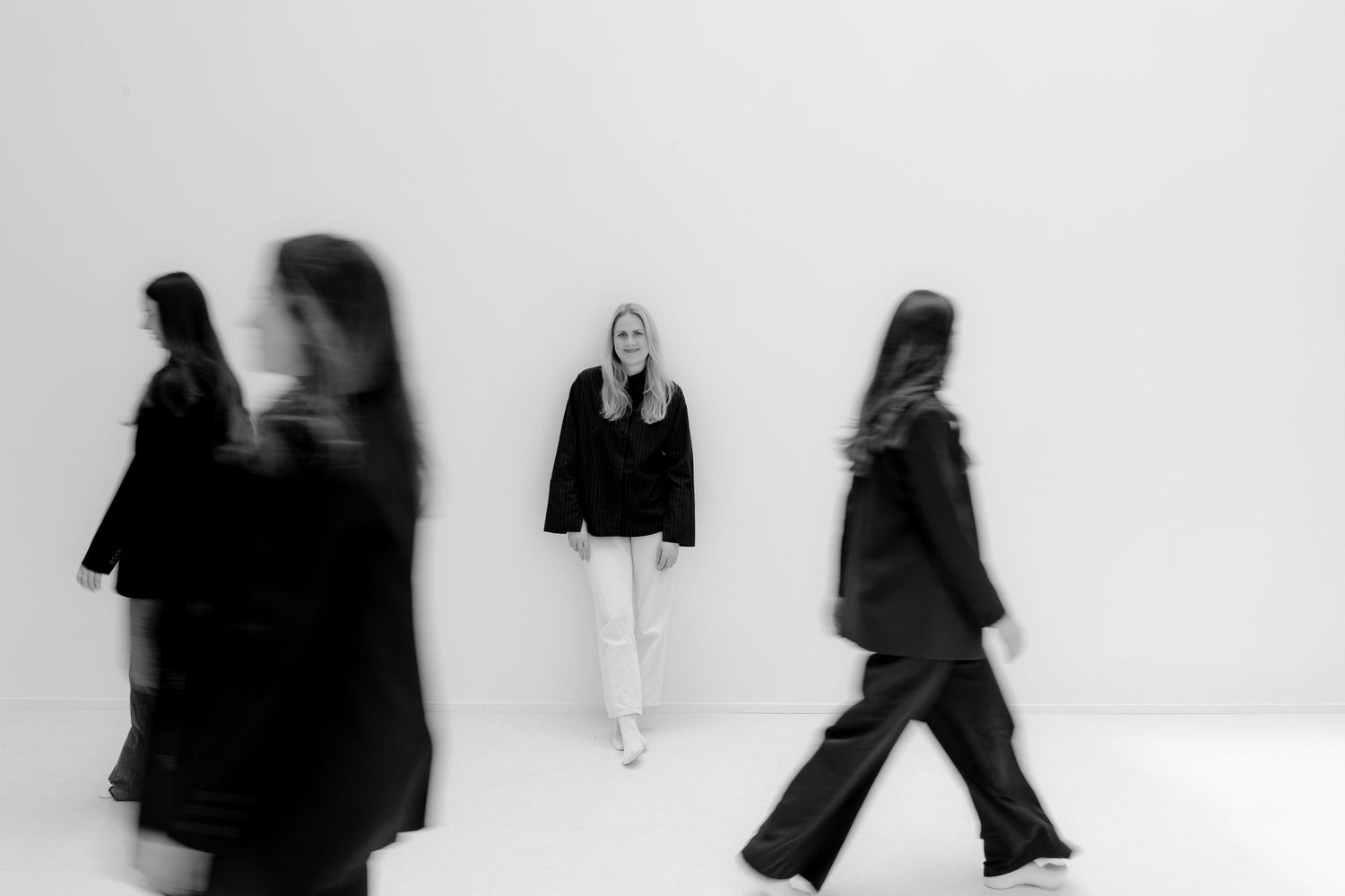 Black and white photo of a blonde woman standing still among blurred moving figures in a minimalist white space.