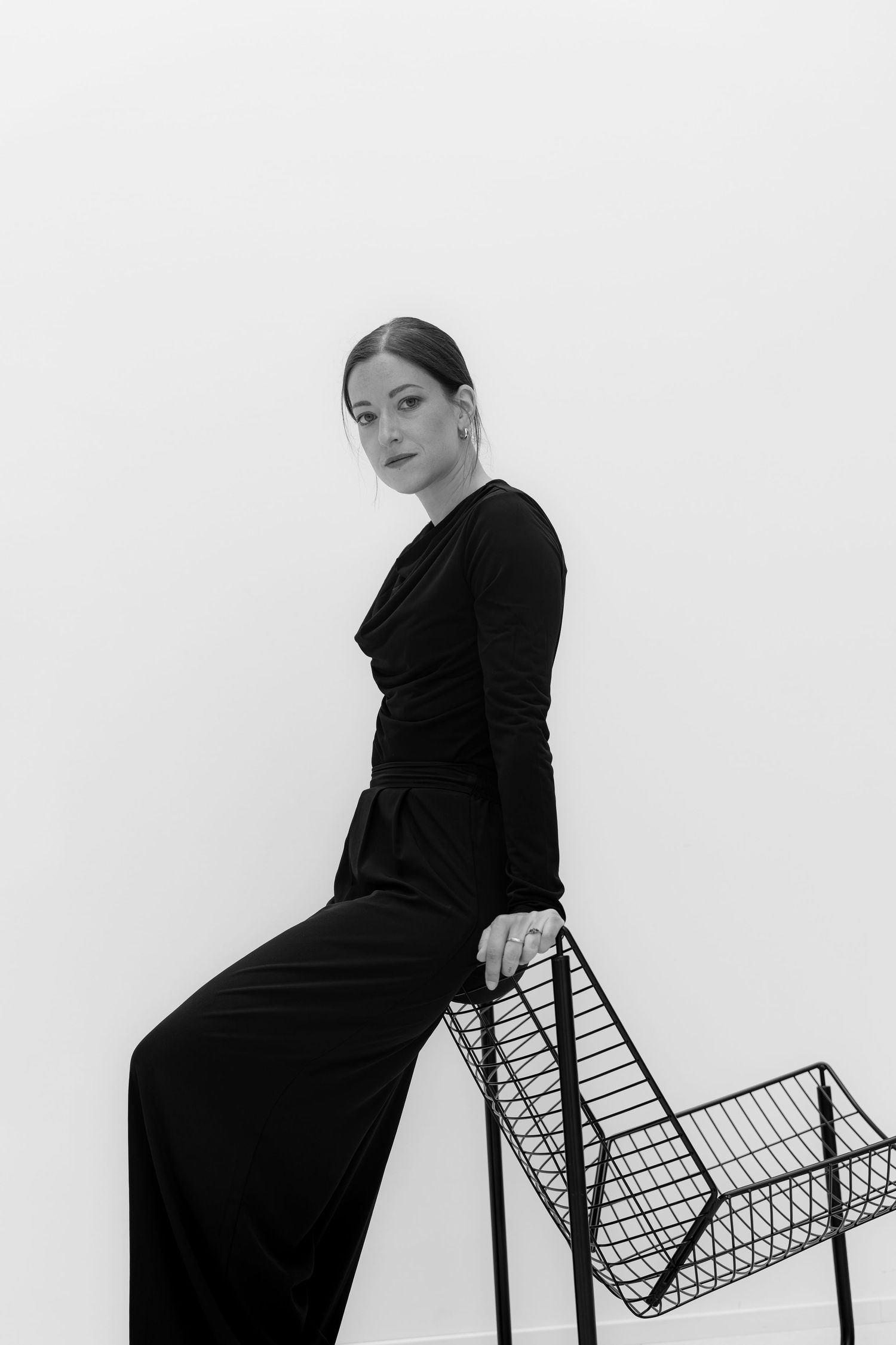 Woman in black dress sitting on wire chair against white background in elegant black and white portrait photo.