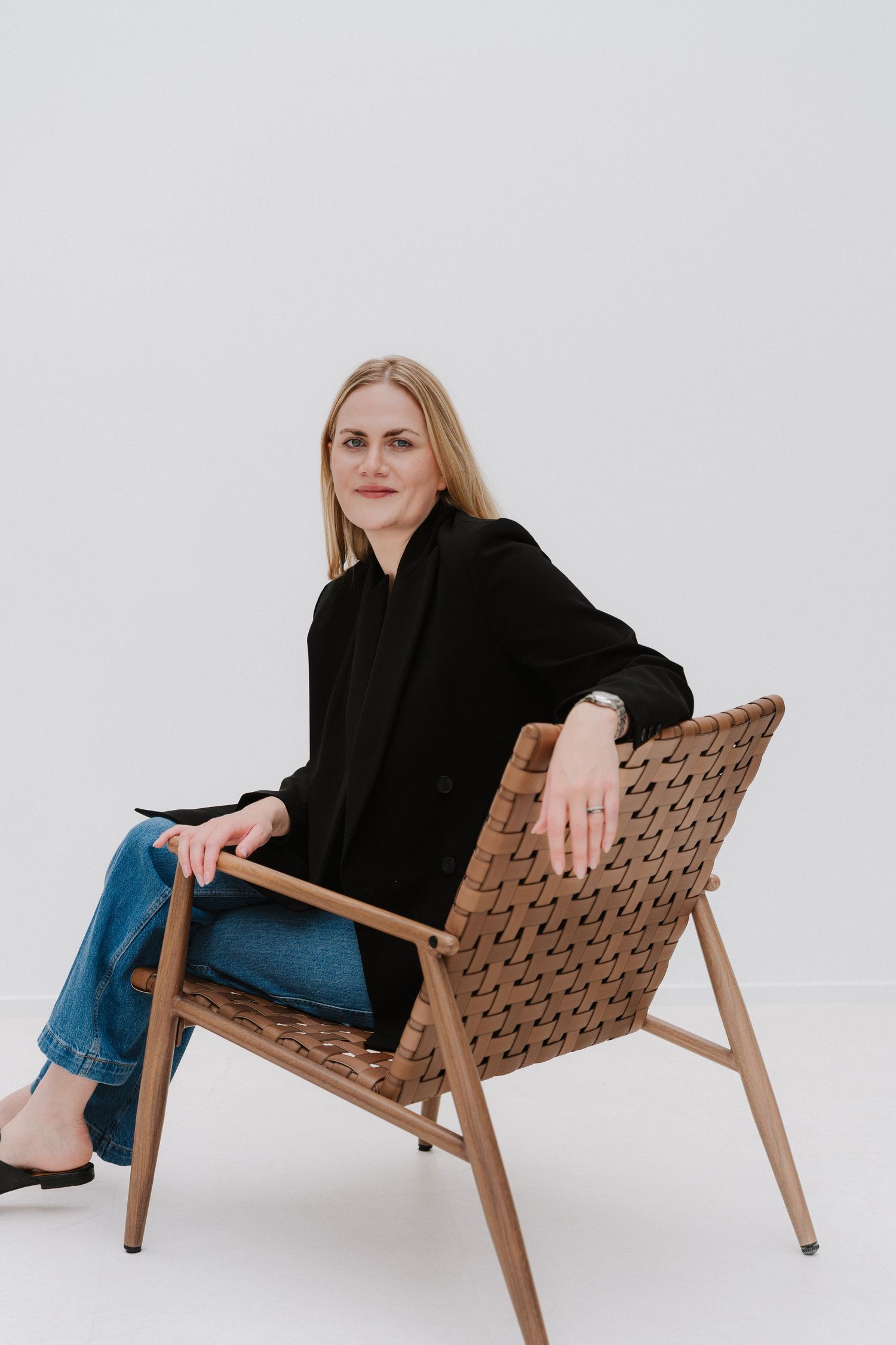 Woman in black blazer and jeans sits relaxed on a woven leather chair against a white background.