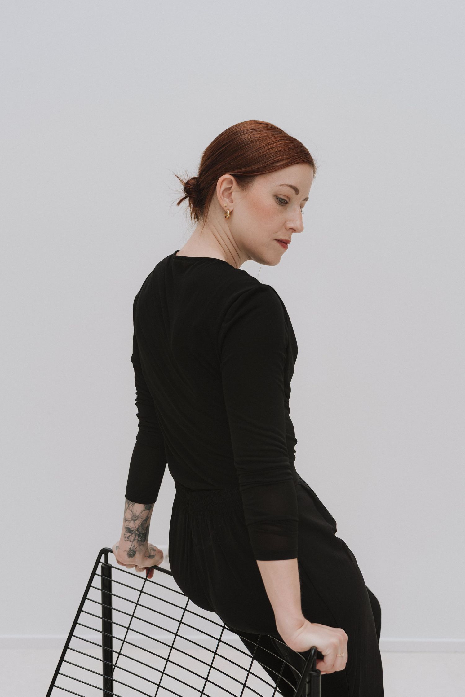 Red-haired woman in black outfit sitting sideways, showing tattoo on arm, holding wire basket on white background.