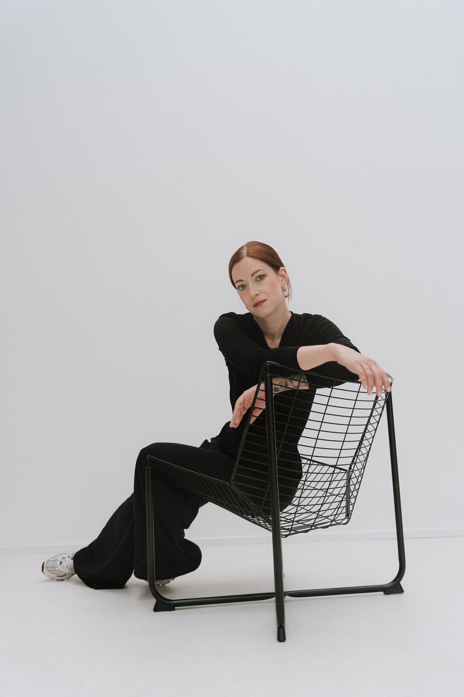 Woman in black outfit sitting on wire chair against white background, posing confidently in casual fashion editorial style.