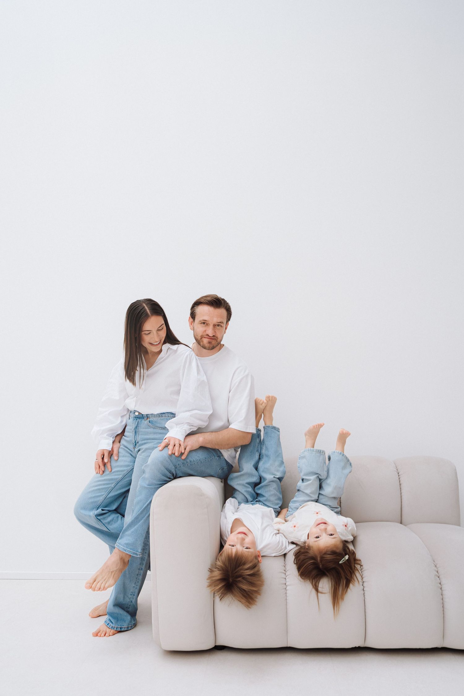 Happy family with two children playing on a white sofa, parents in white tops and jeans, kids hanging upside down.