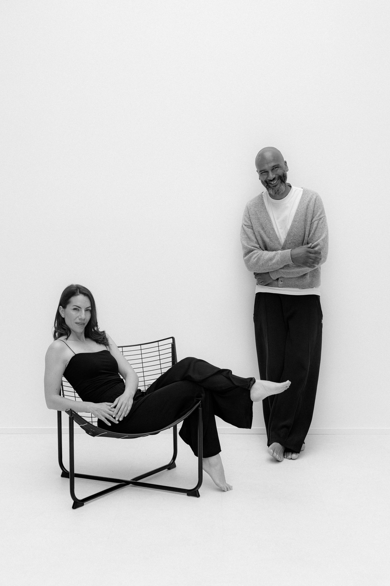 Black and white photo of a pregnant woman sitting in a chair beside a bald man with arms crossed against a white wall.