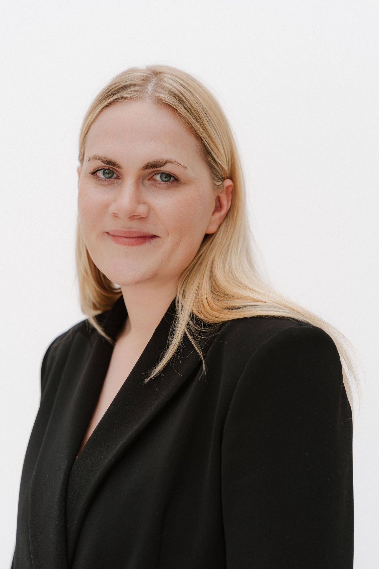 Professional blonde woman in black blazer poses confidently against white background for business headshot.