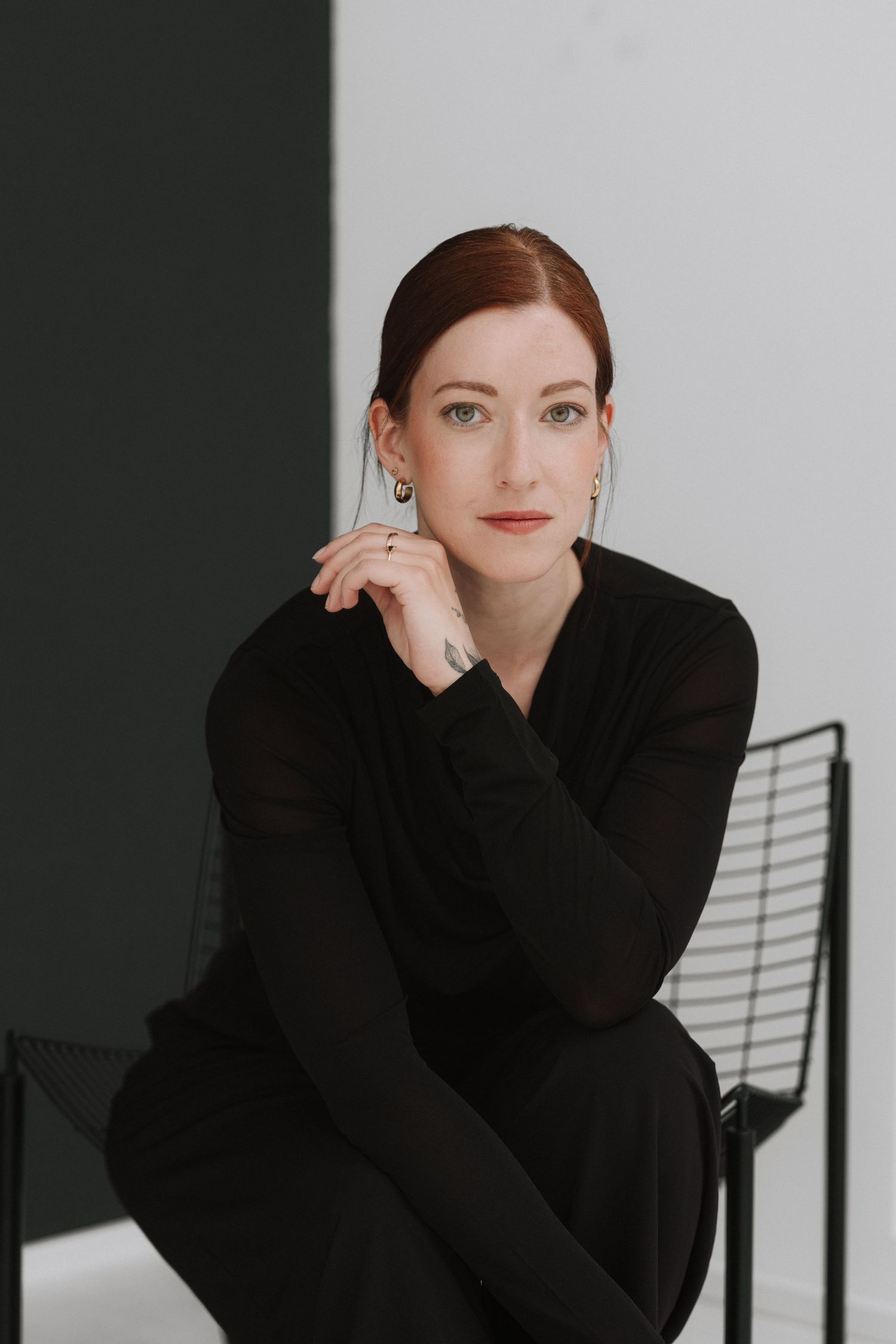 Woman in black outfit sitting on metal chair against green and white wall, with red hair and silver earrings.