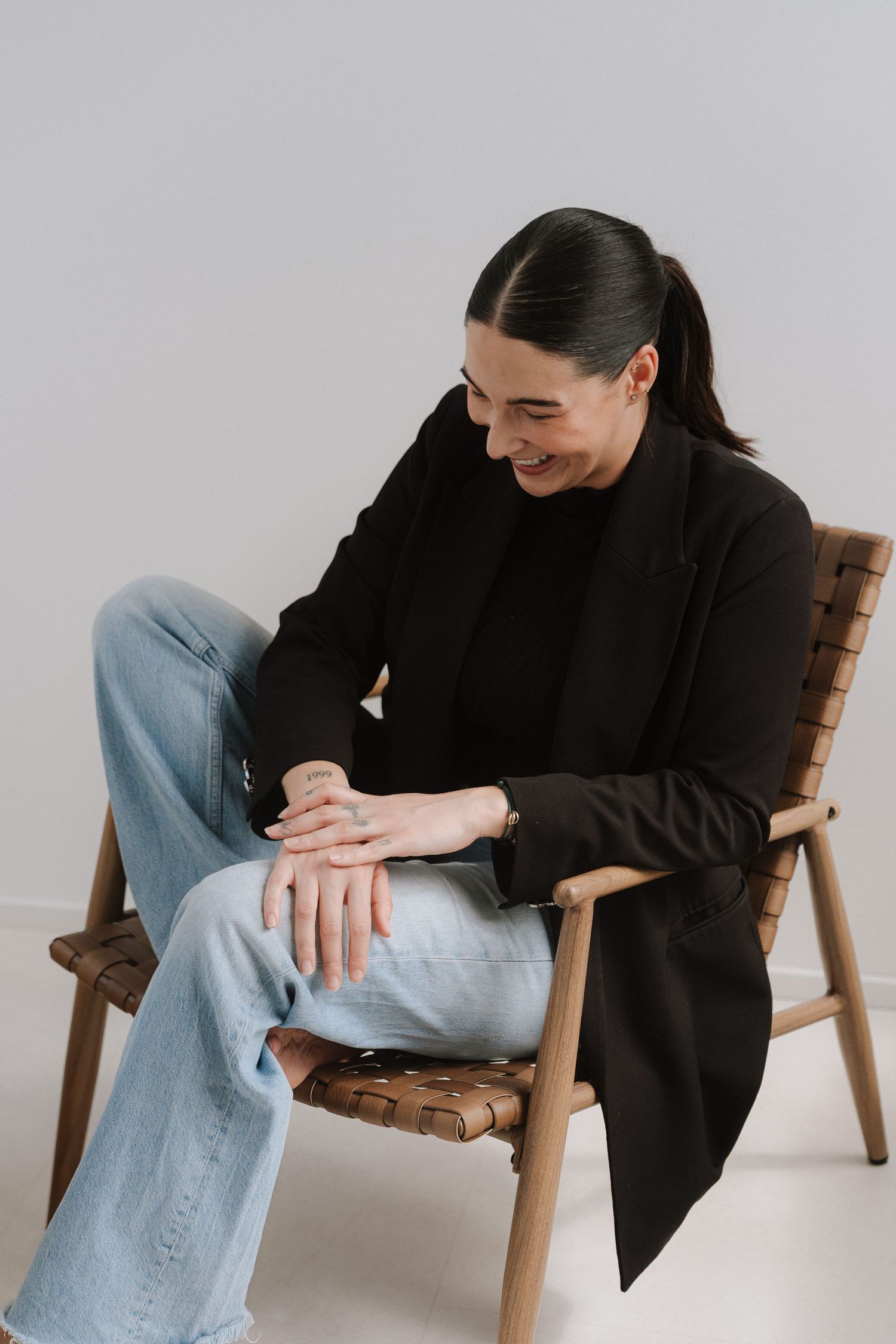 Smiling person in black blazer and jeans sitting relaxed in a wooden chair against a light gray background.