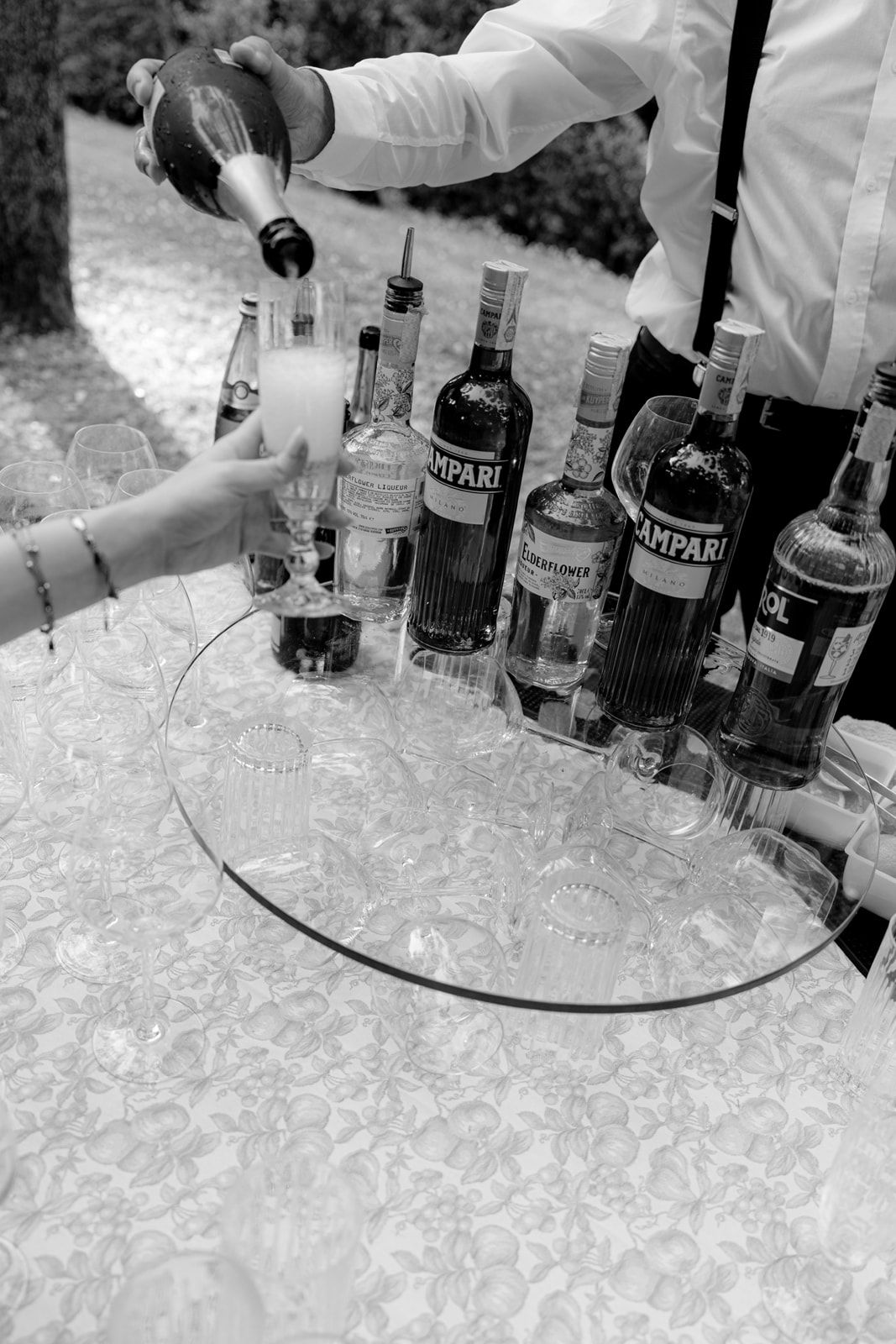 Black and white photo of bartender pouring champagne at event bar with Campari bottles and glasses on table.