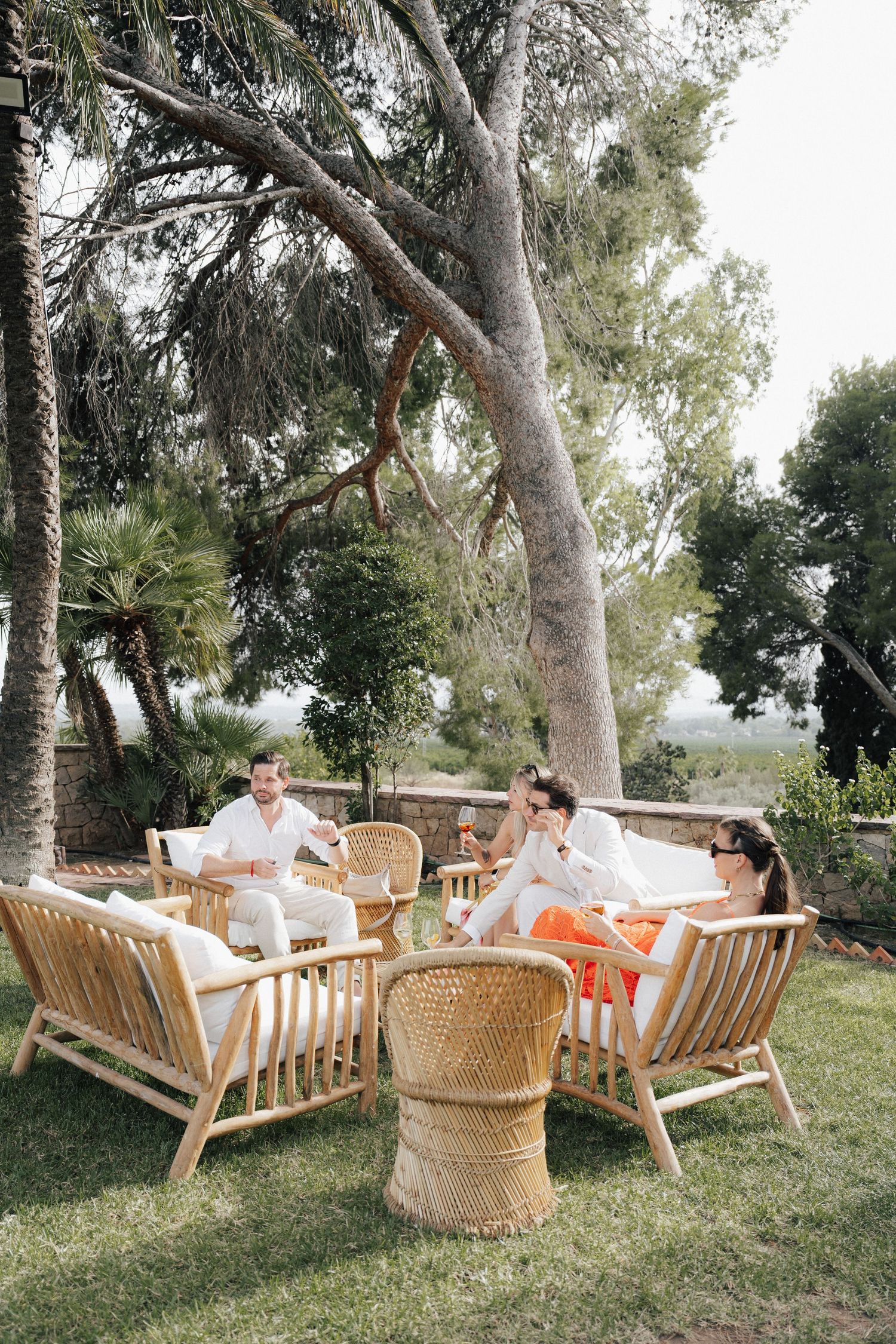 Outdoor lounge area with wicker furniture and cushions arranged under tall trees.