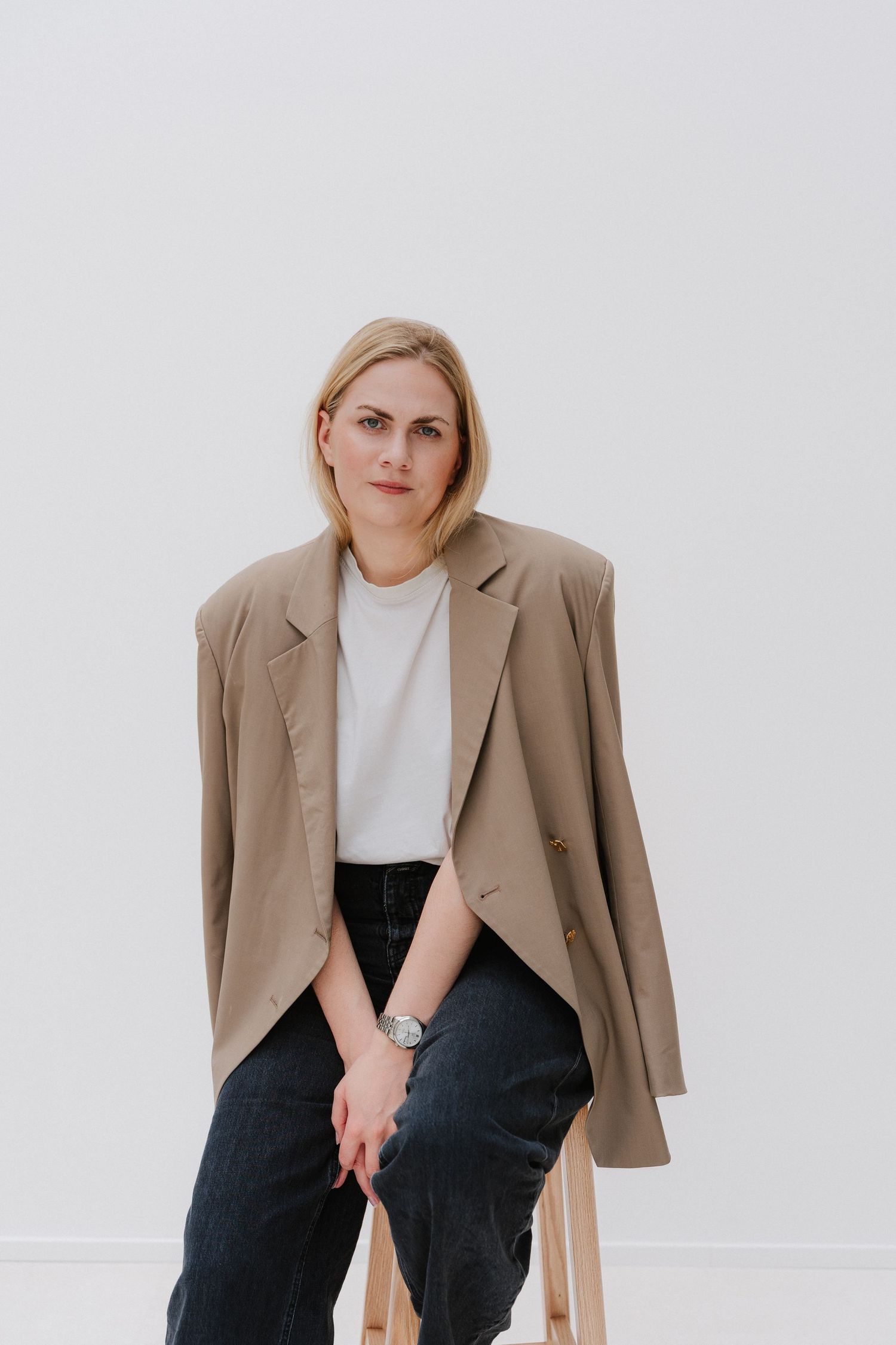 Blonde woman in beige blazer and white tee sits on stool against white wall, wearing a silver watch.