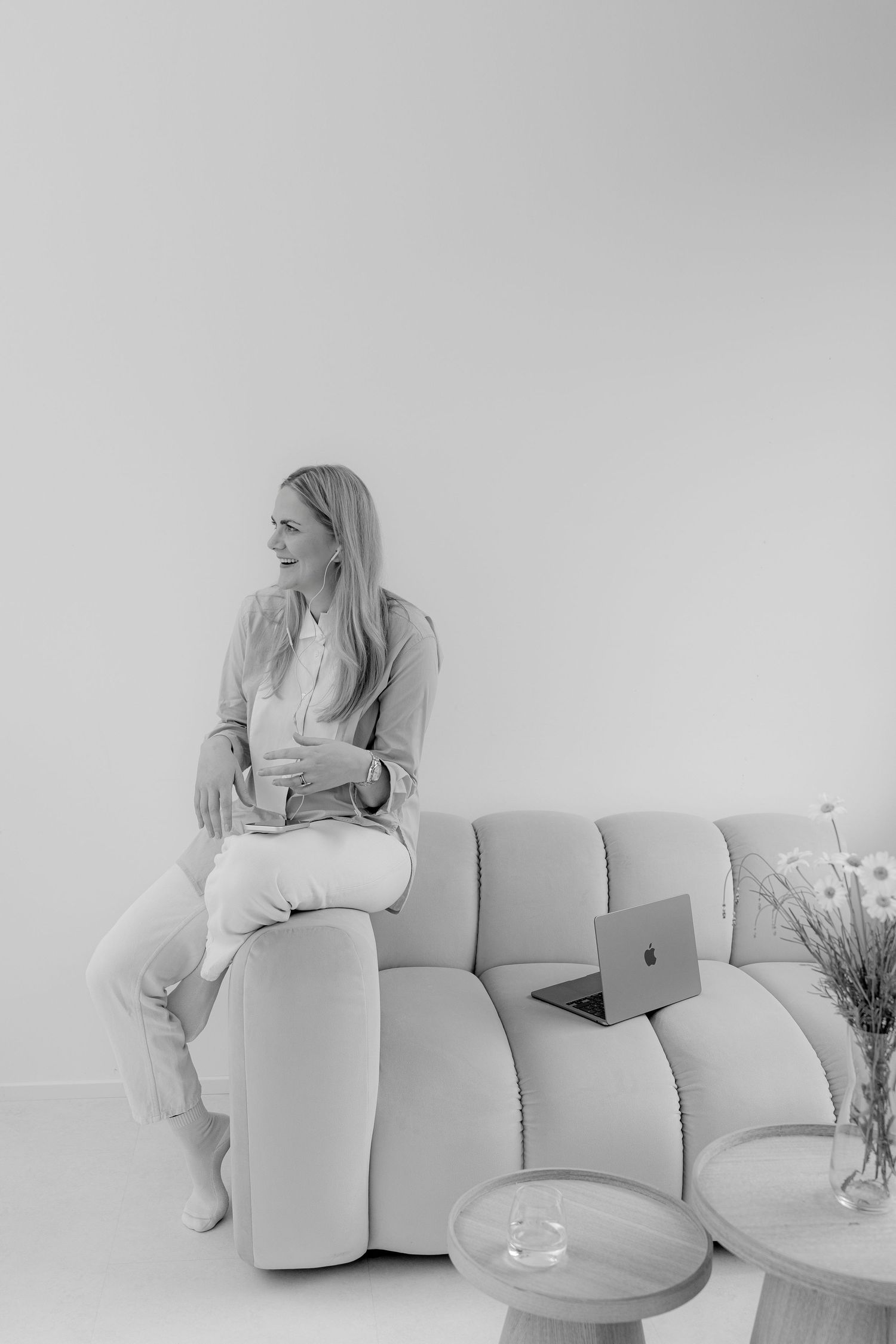 Woman sitting on modern sofa with laptop, smiling thoughtfully in bright minimalist room.