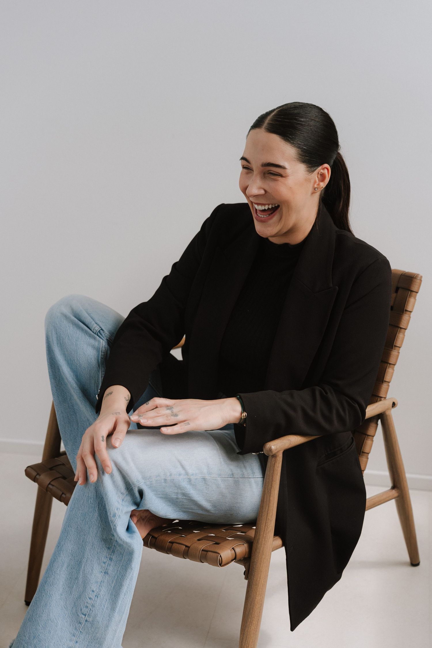 Woman in black blazer and jeans laughing while seated in a wooden leather chair against a white background.