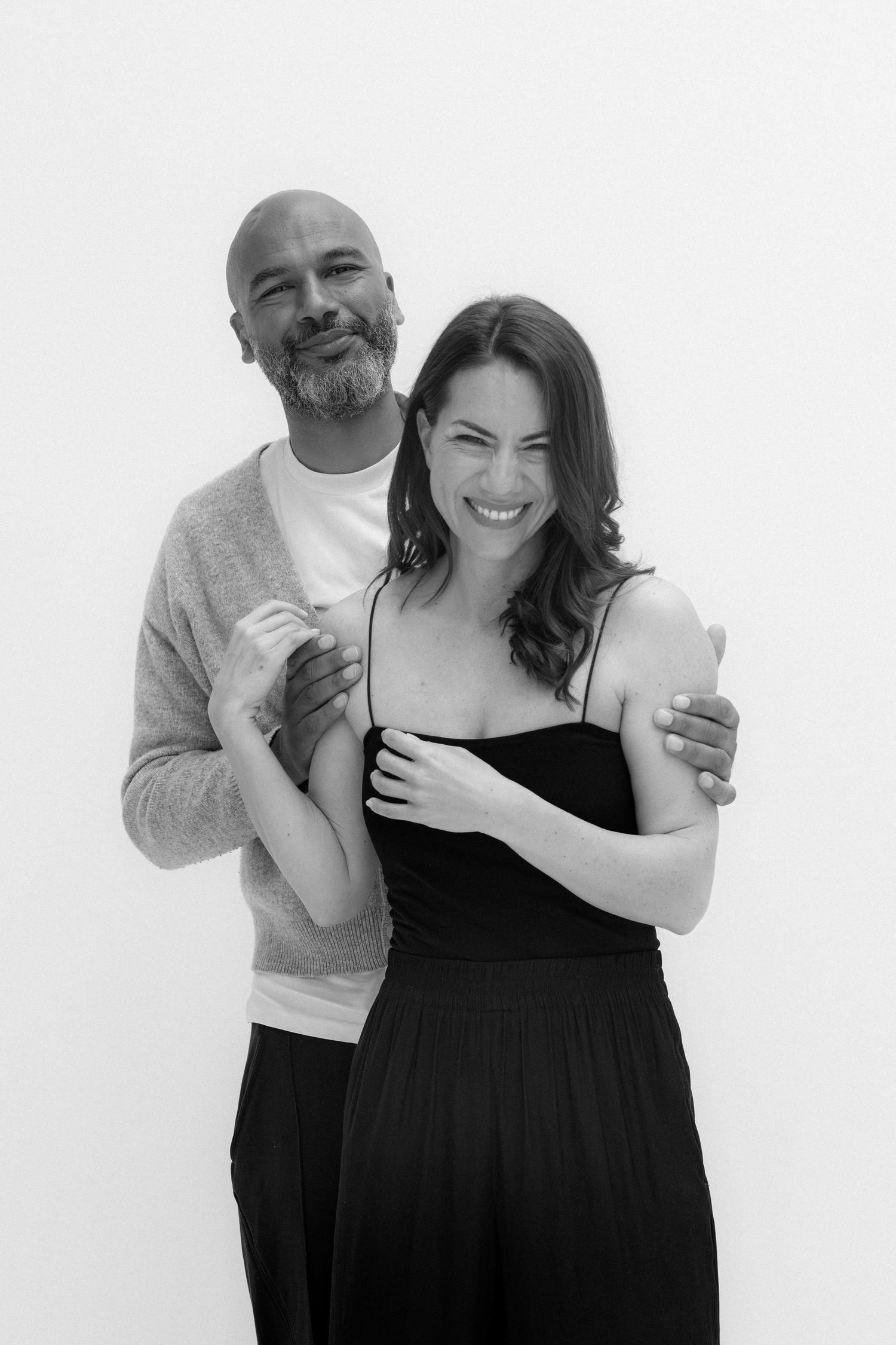 Black and white portrait of a bald man and smiling woman in black dress embracing against white background.