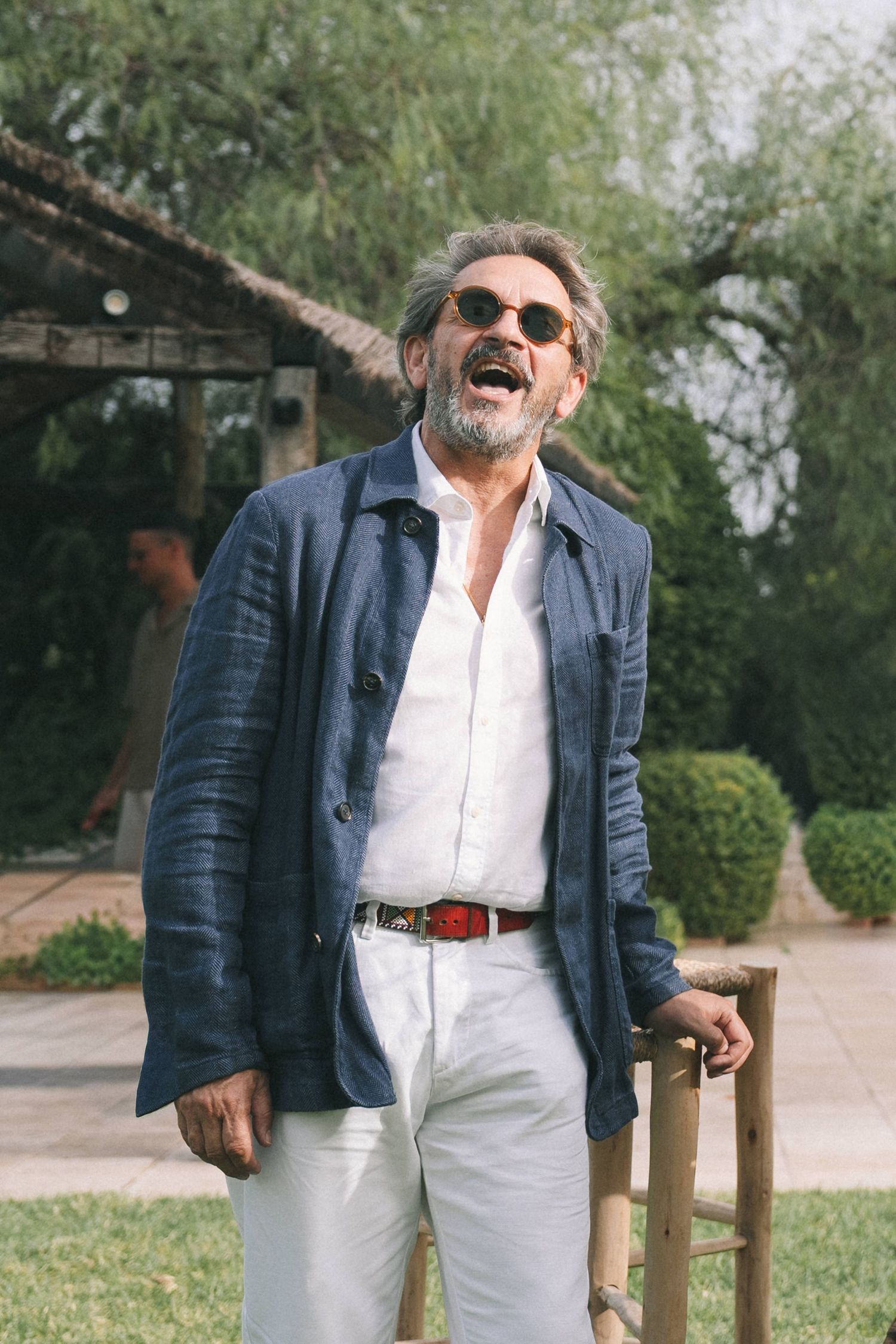 Guest laughing joyfully at outdoor garden party wearing navy blazer and sunglasses.