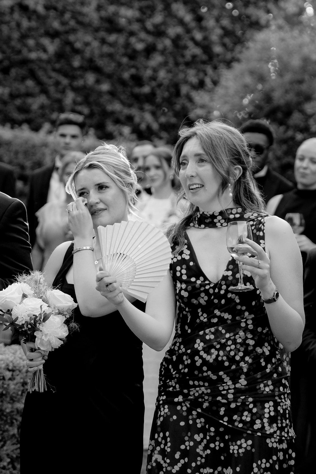 Black and white photo of two women at a wedding, one holding a fan and bouquet, the other holding a wine glass.
