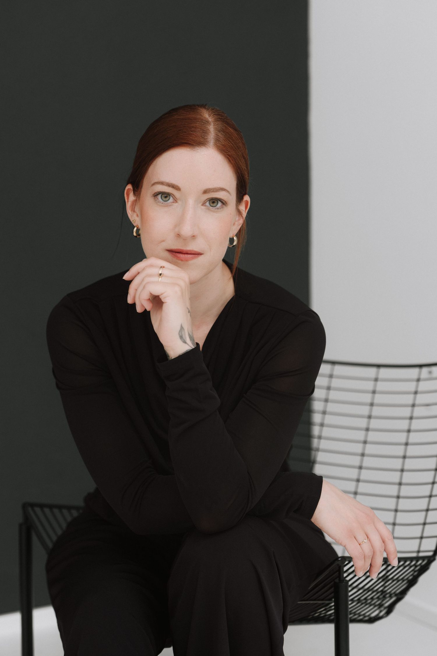 A woman with red hair in black outfit sits on a wire chair, posing thoughtfully with hand on chin.