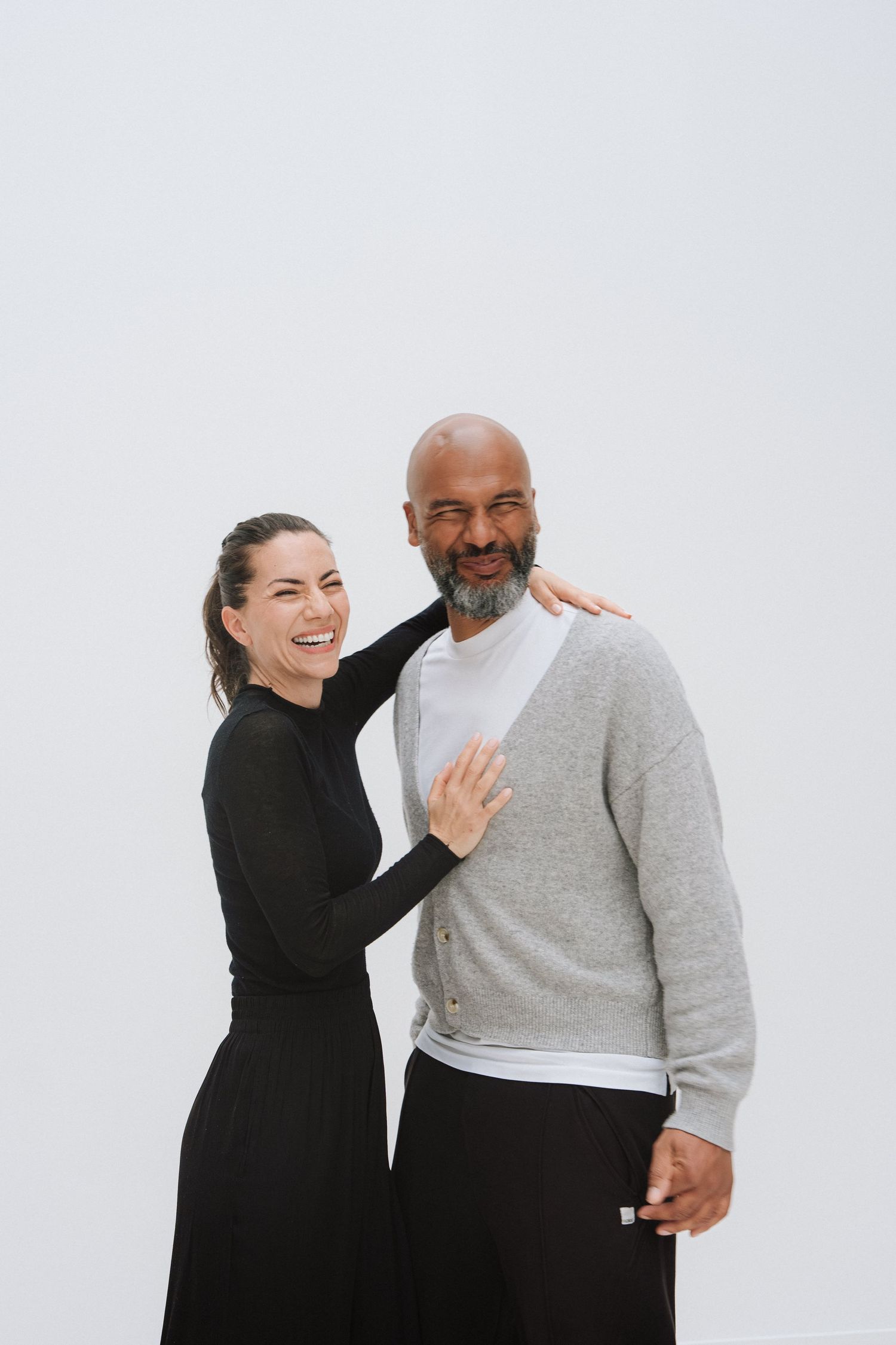A smiling interracial couple posing together, woman in black dress with arm around bald bearded man in gray cardigan.