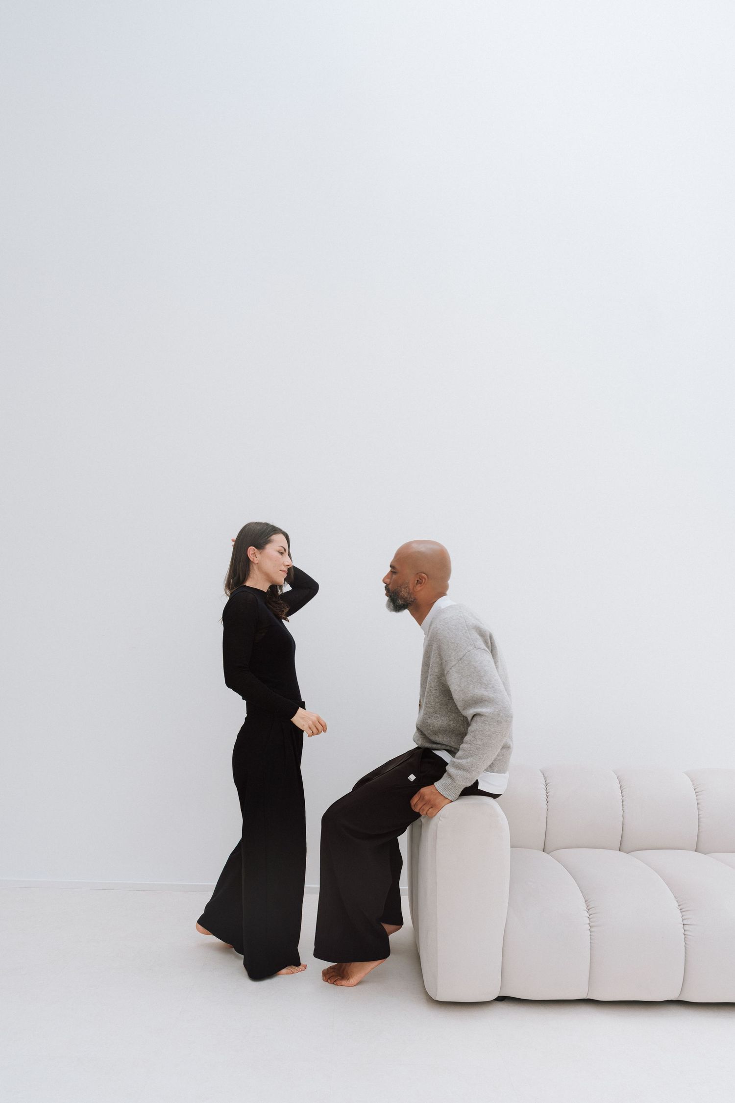 A man and woman in a minimalist white room, she in black outfit standing, he in gray sitting on a white sofa.
