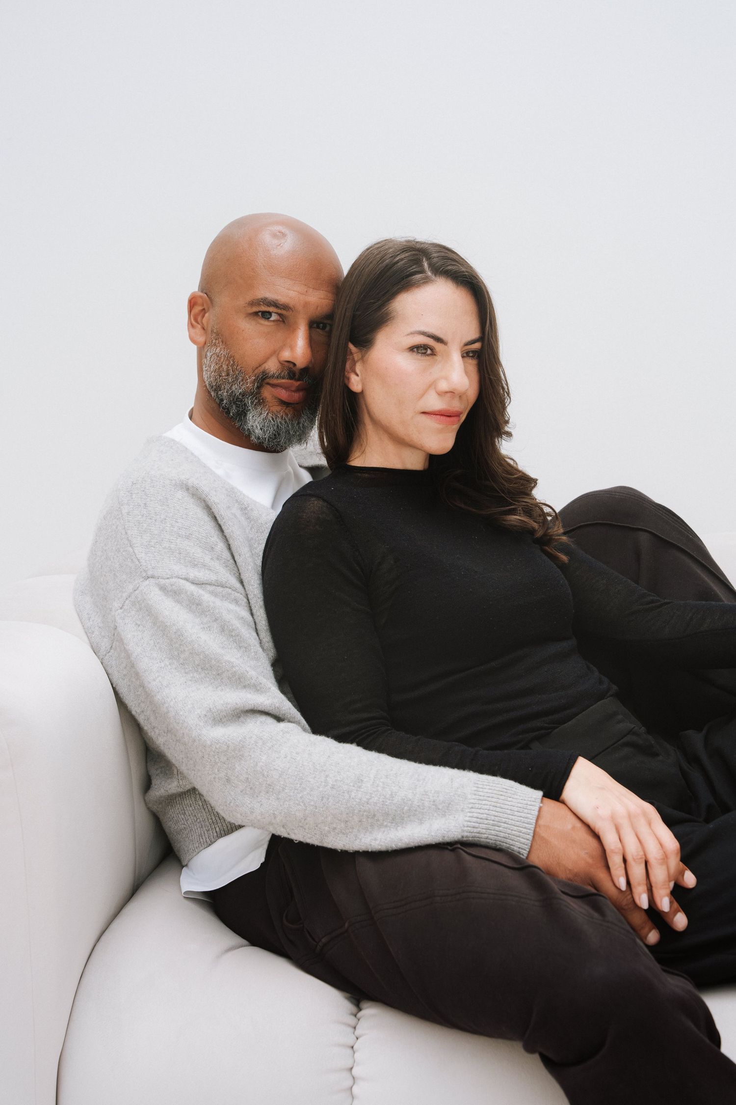 A couple sits together on a white sofa, man in gray sweater and woman in black outfit, posing for a portrait.