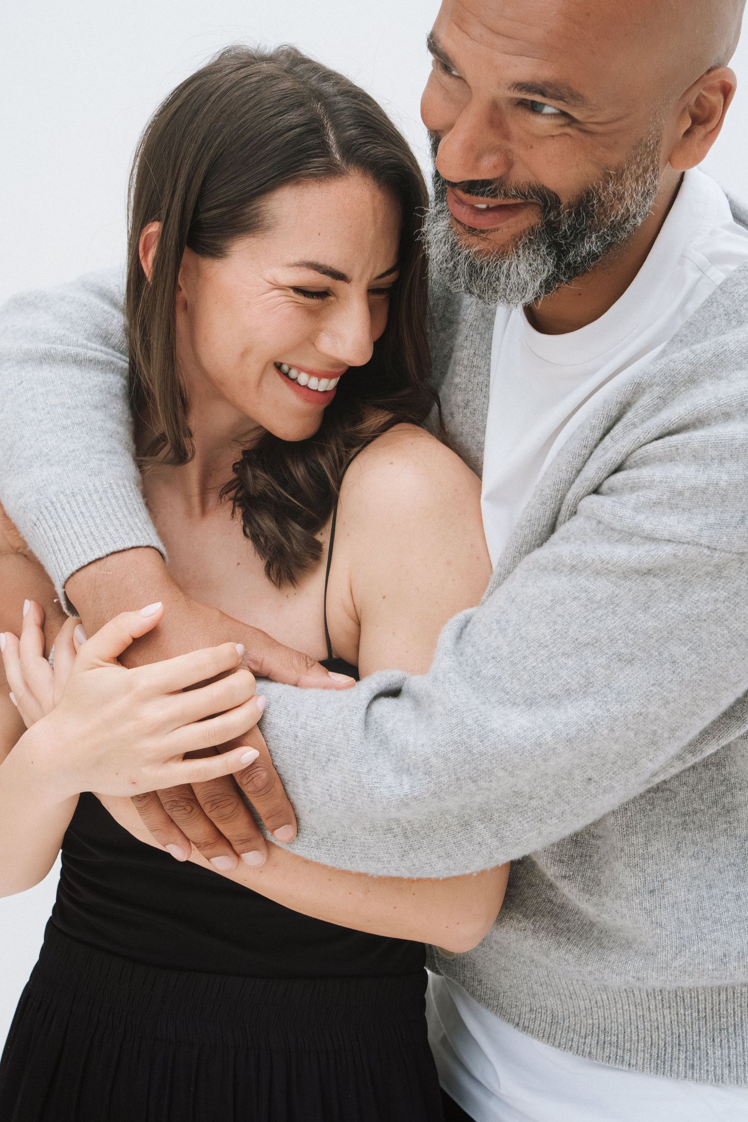 A smiling couple embracing warmly, man with beard in gray sweater hugging a laughing brunette woman in black skirt.