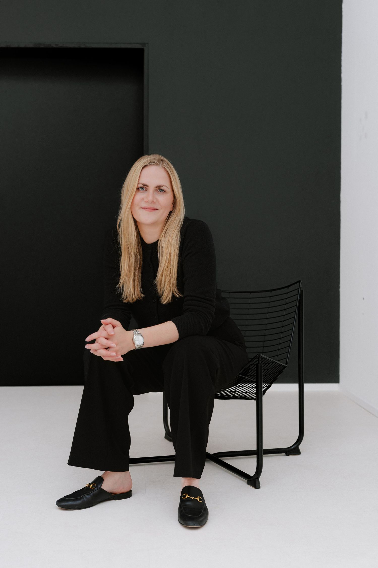 Woman in black outfit sitting on a modern black chair against a dark green and white minimalist background.
