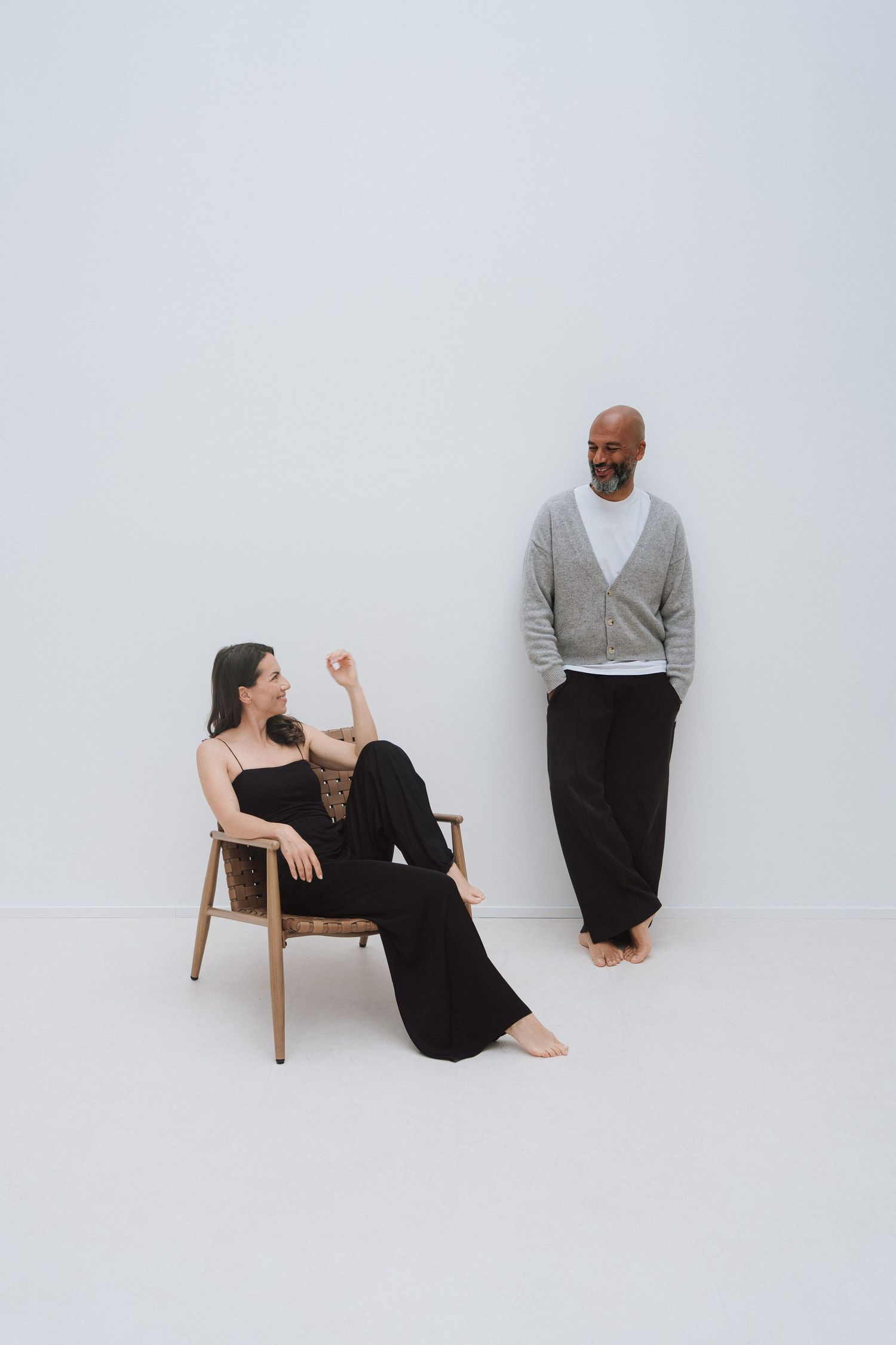 A woman in black sits in a wooden chair while a bald man in gray cardigan stands against a white wall.