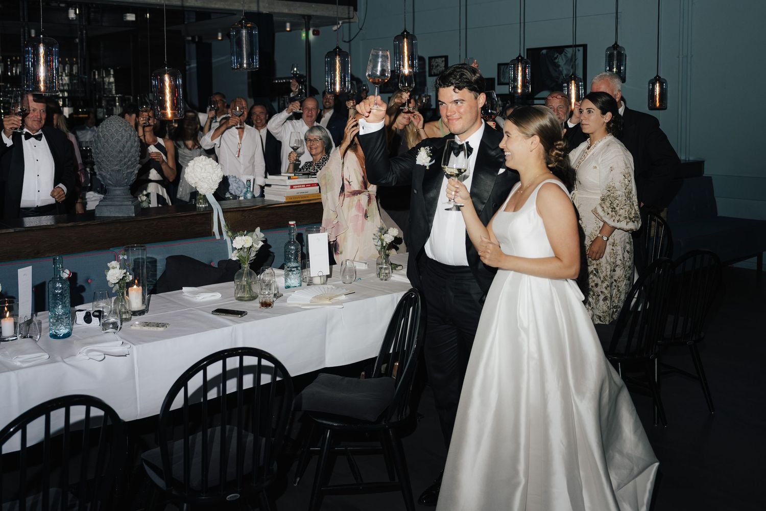 Wedding reception guests seated at tables in a dimly lit venue with elegant decor and white tablecloths.