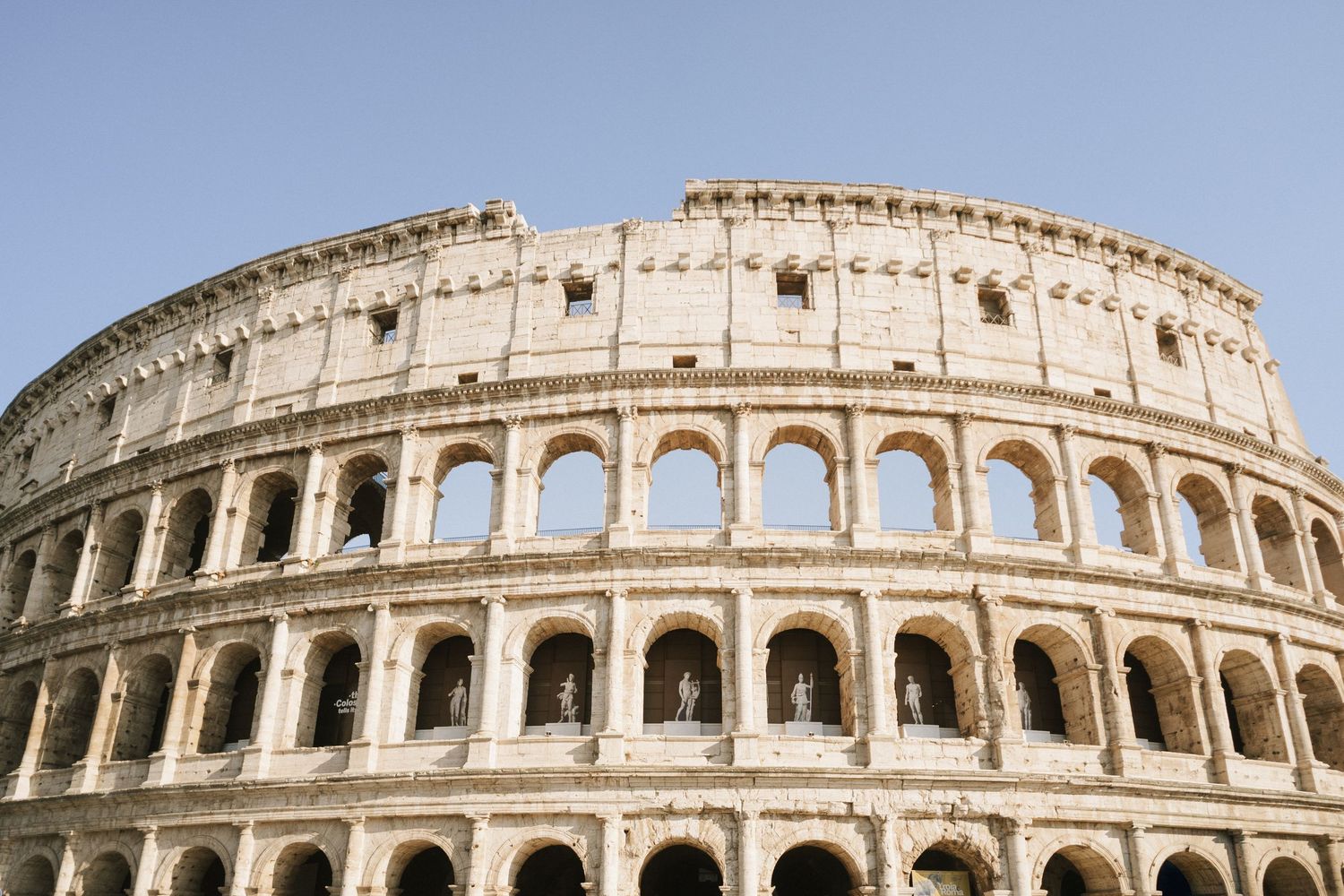 Ancient Roman Colosseum exterior with iconic arched facades and weathered stone walls against a clear blue sky.