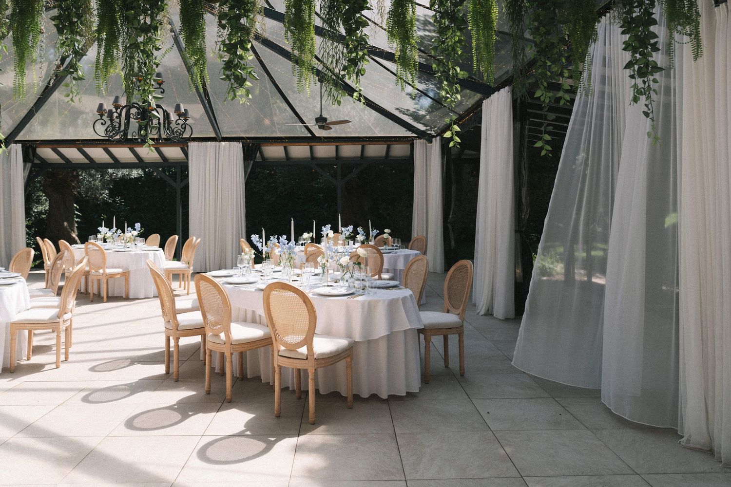Elegant outdoor wedding reception under a glass pavilion with white draped tables, gold chairs, and lush greenery.