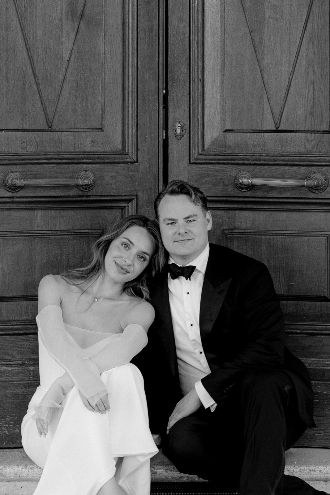 Black and white photo of a bride and groom sitting together in front of elegant wooden doors on their wedding day.