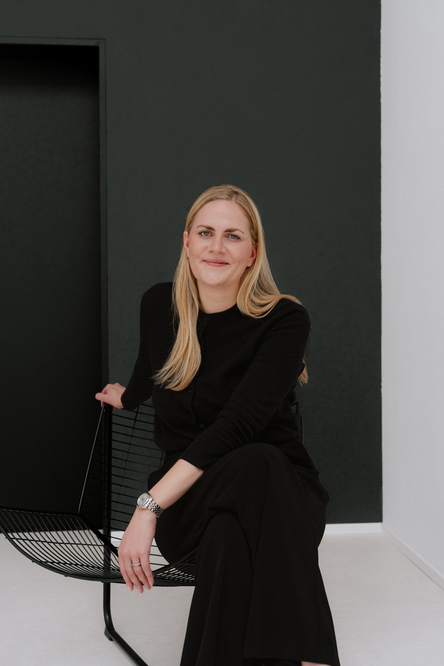 Blonde woman in black outfit sitting on a modern wire chair against a dark green wall, smiling confidently.