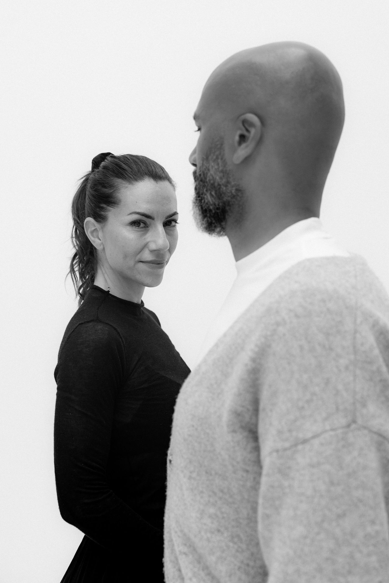 Black and white portrait of a woman in black turtleneck and bald bearded man in light sweater facing each other.