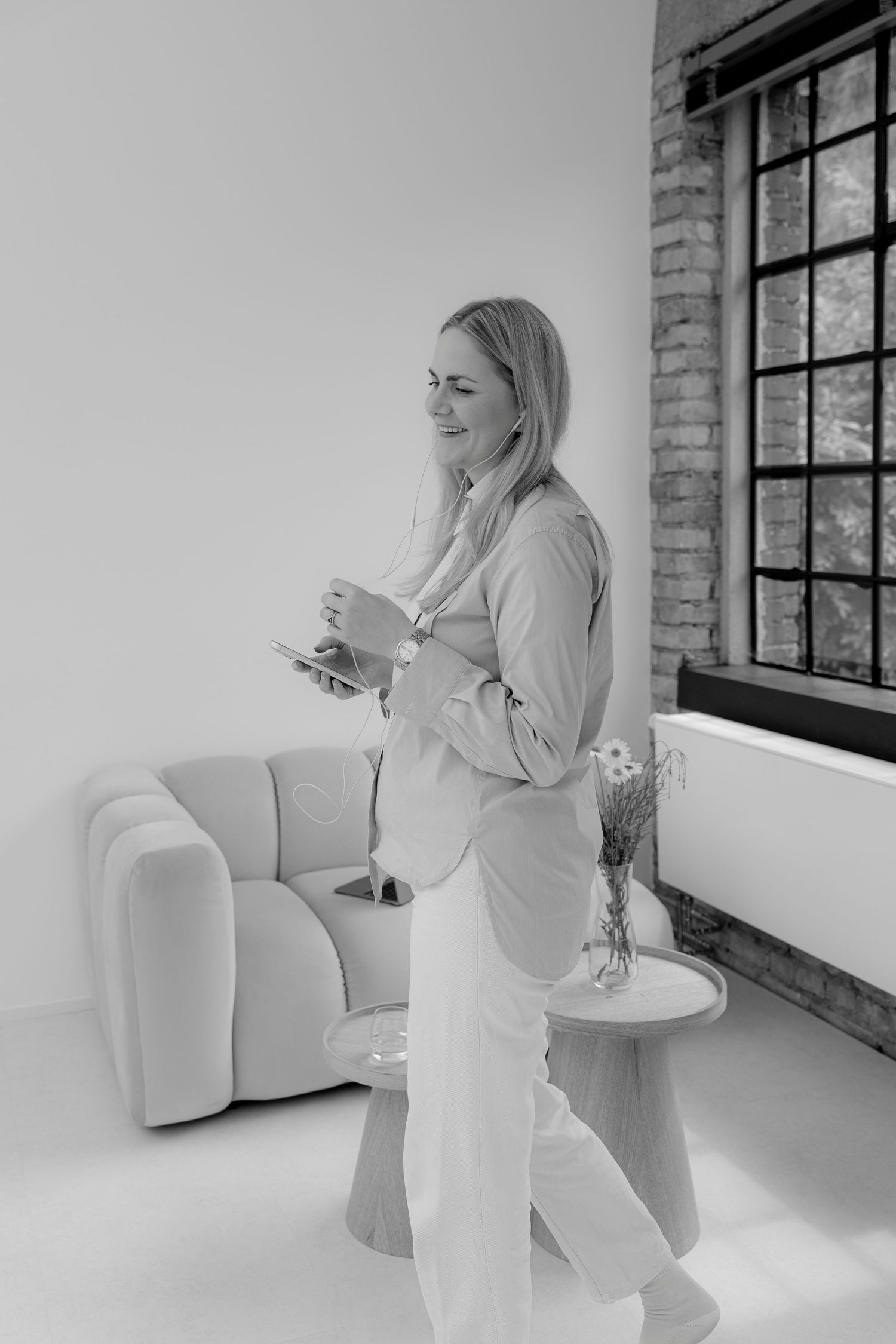 Black and white photo of a smiling blonde woman in a blazer holding a tablet, standing near a window in a modern interior.