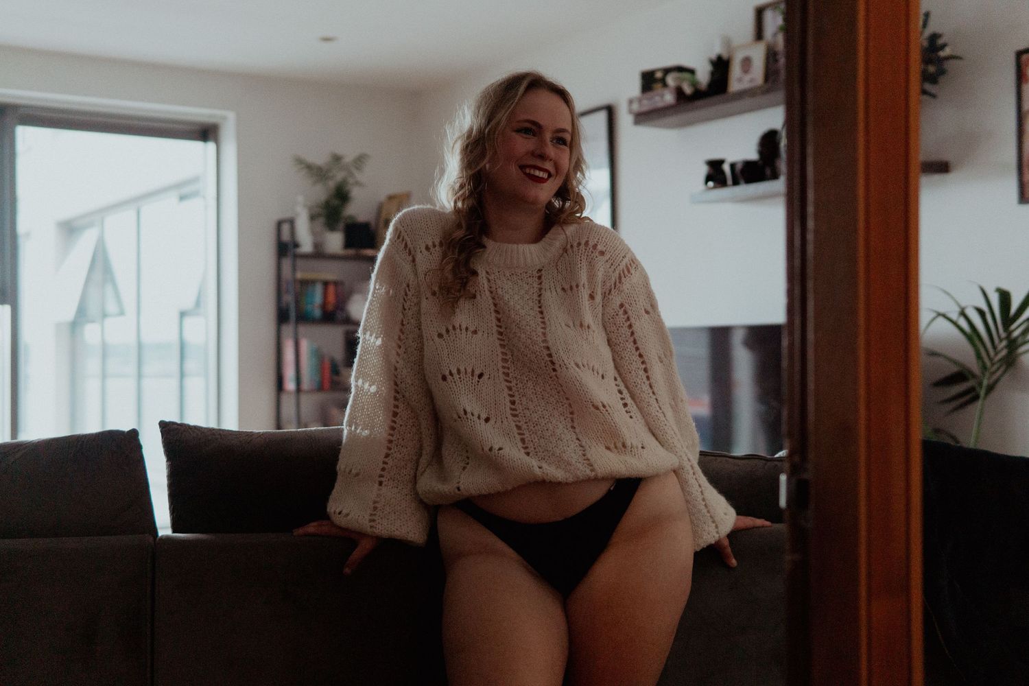 Candid boudoir moment of woman laughing in oversized sweater and black underwear, leaning on the back of a sofa.