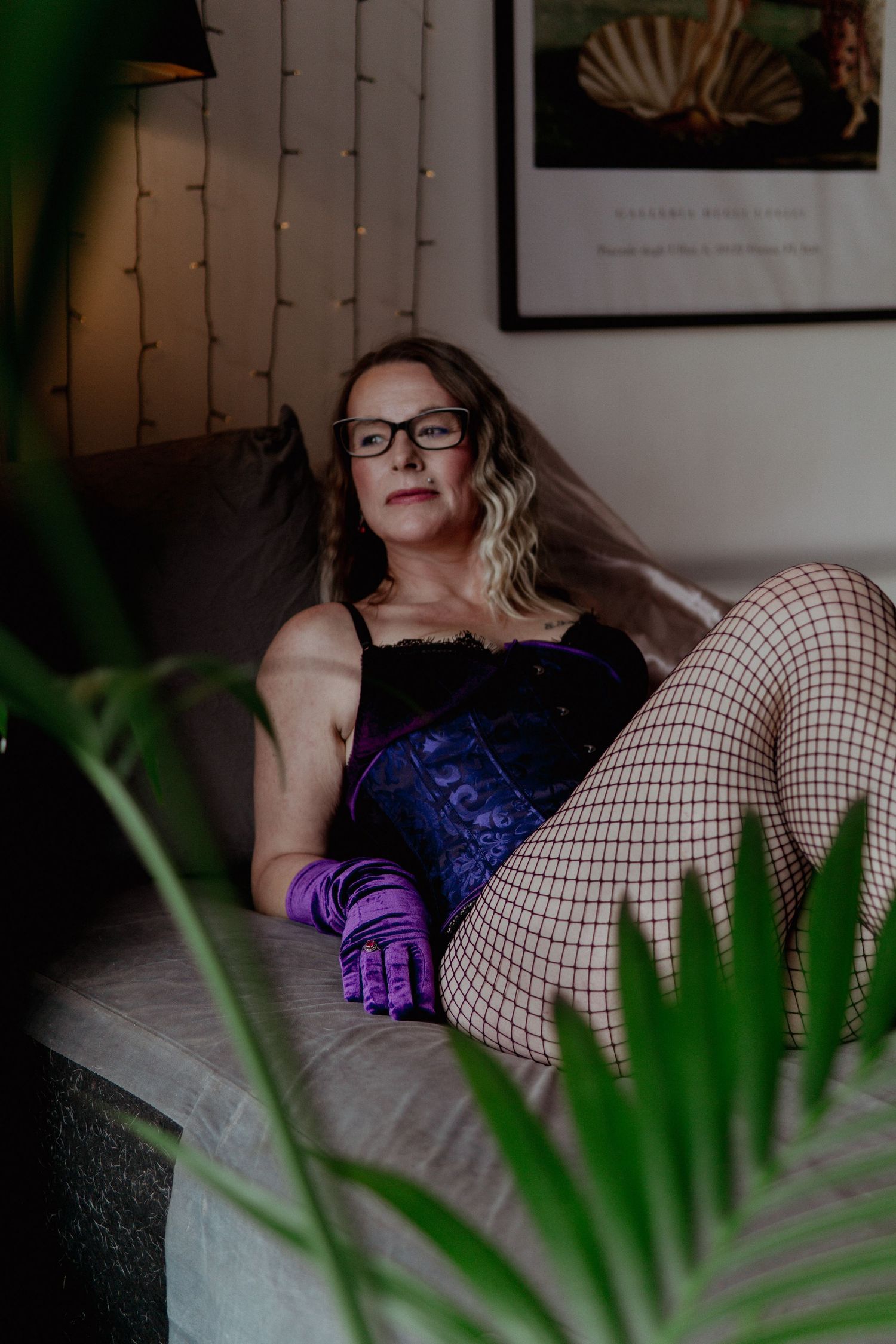 A woman in blue corset and fishnet stockings reclines on a couch surrounded by indoor plants. Liverpool boudoir photography