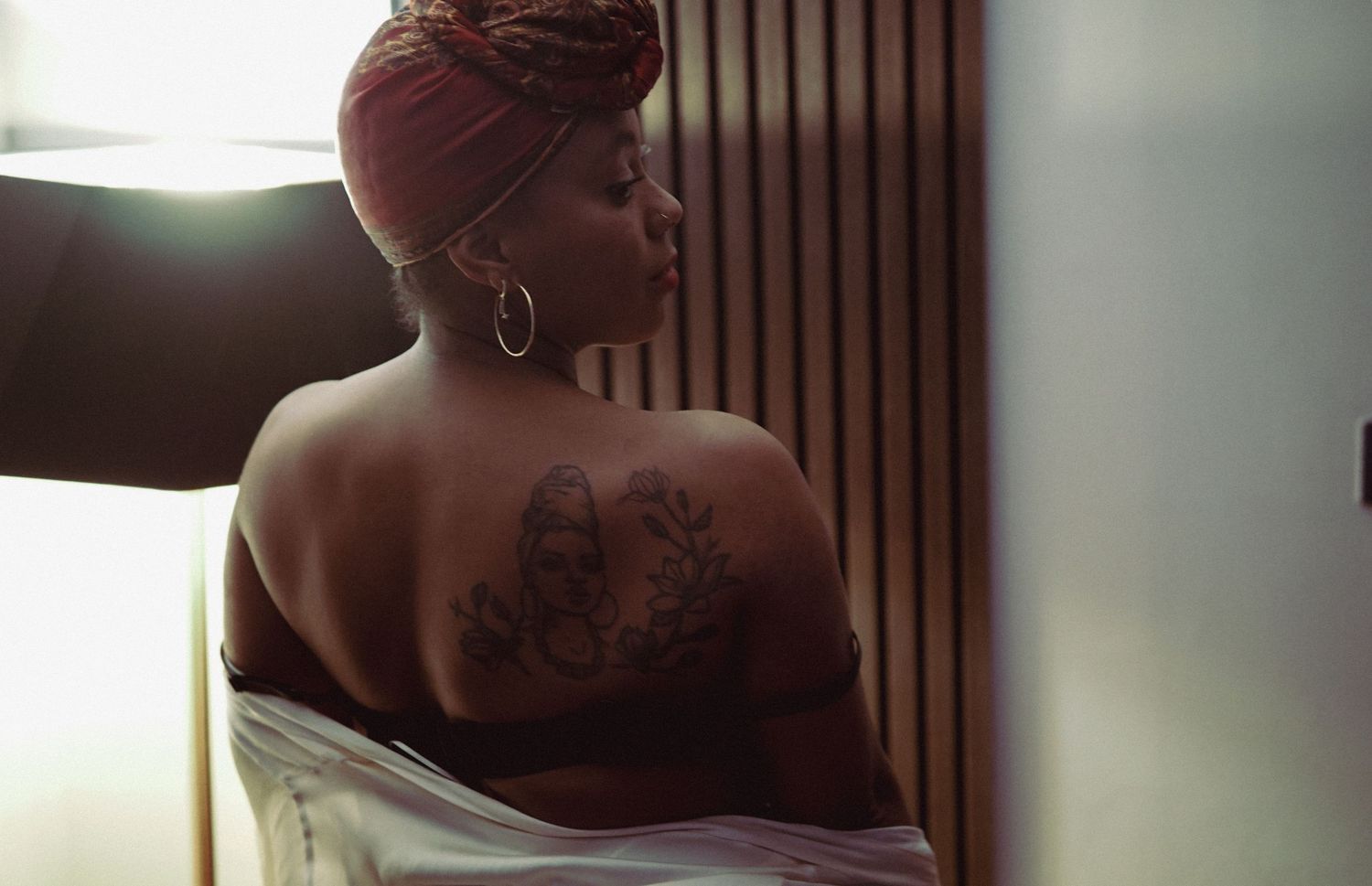 A woman with a head wrap reveals an intricate back tattoo while draped in a white cloth. Liverpool boudoir photography