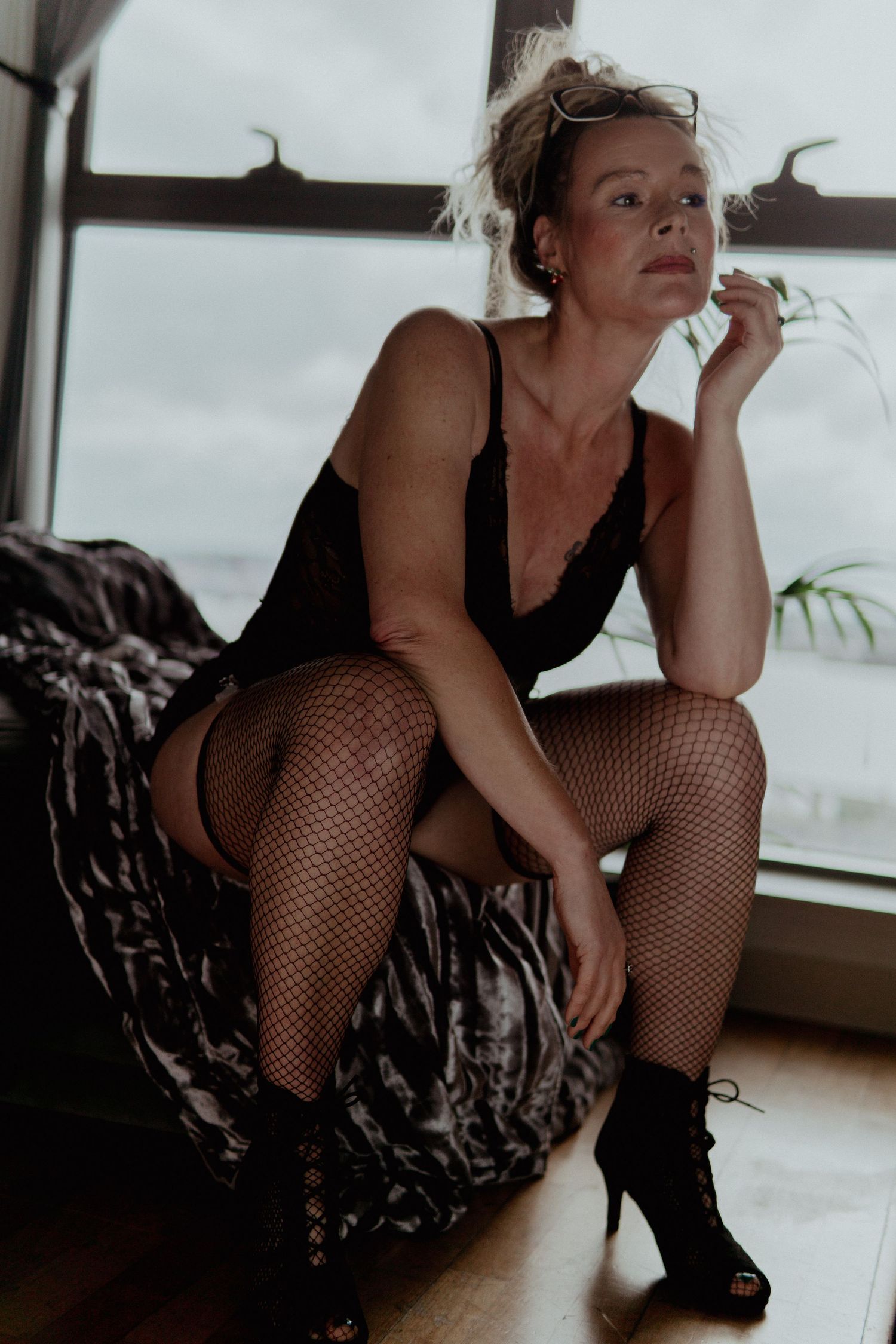 Woman in black lingerie and fishnet tights posing on a bed near a window. Liverpool boudoir photography
