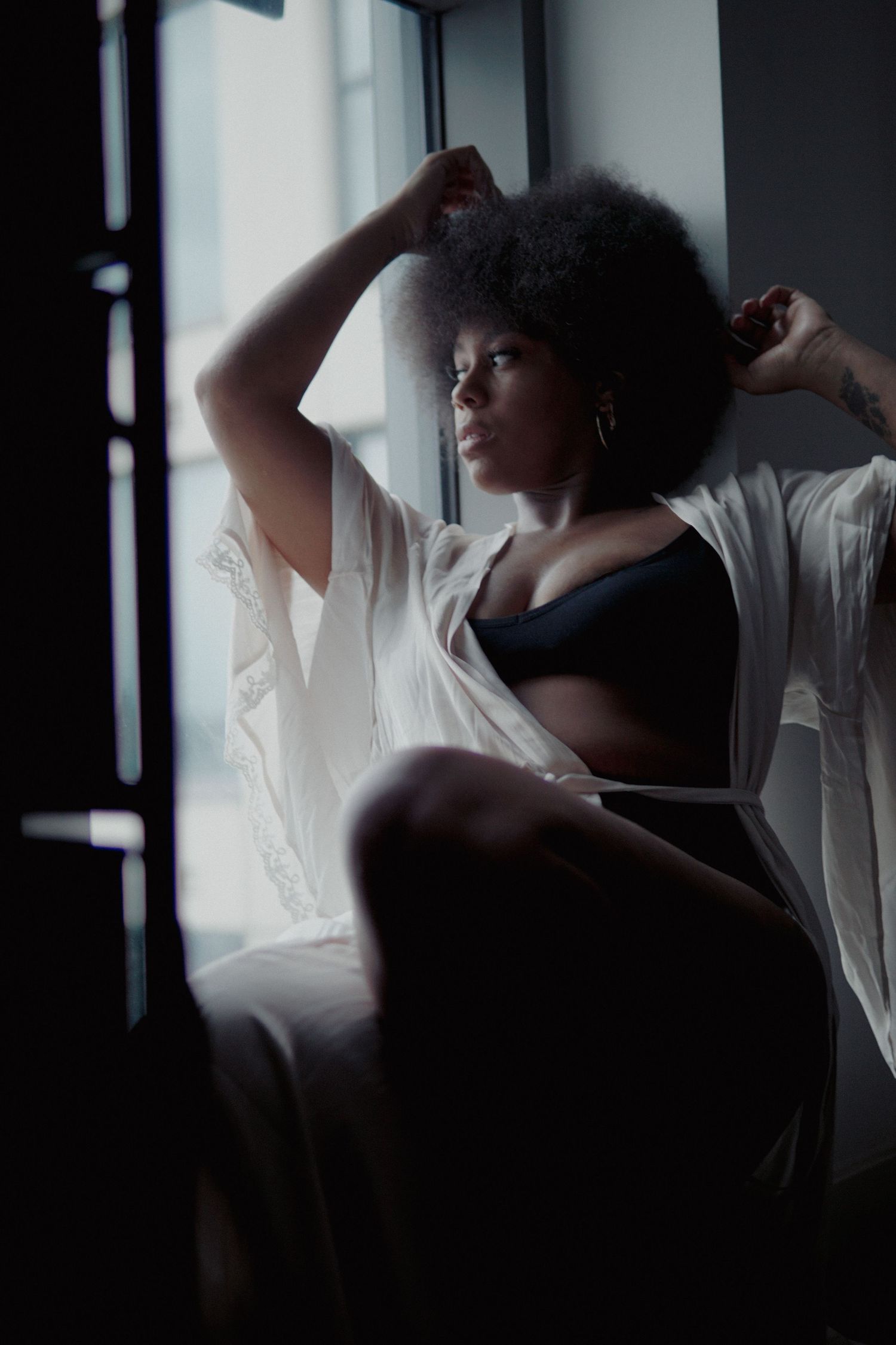 Woman with afro in black underwear and white robe sitting on window seal and looking out of window Boudoir photography UK Liverpool