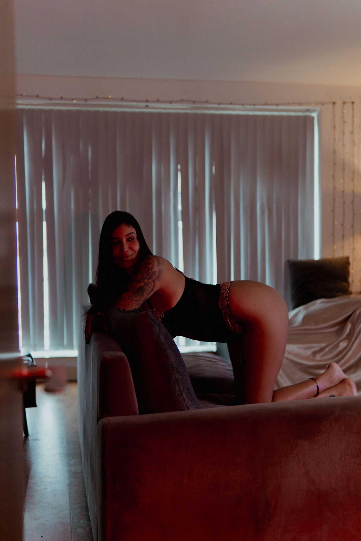 Woman in black outfit posing on a velvet sofa in a dimly lit room with vertical blinds. Liverpool boudoir photography