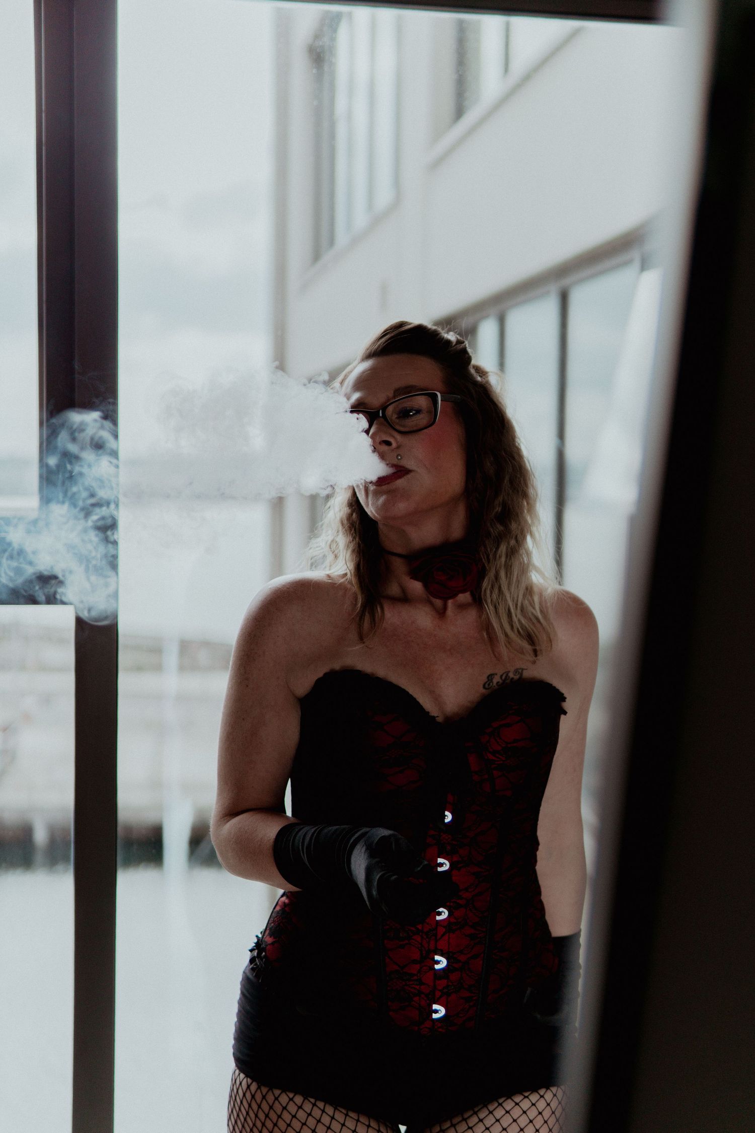 Woman in red corset and fishnet stockings smoking near a window. Liverpool boudoir photography