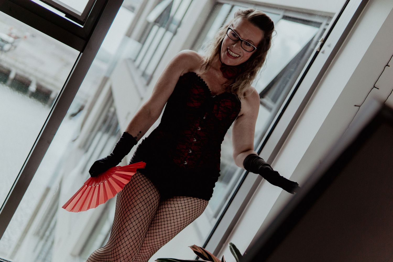 Smiling woman in black corset with fishnet stockings holding red fan near large windows. Liverpool boudoir photography