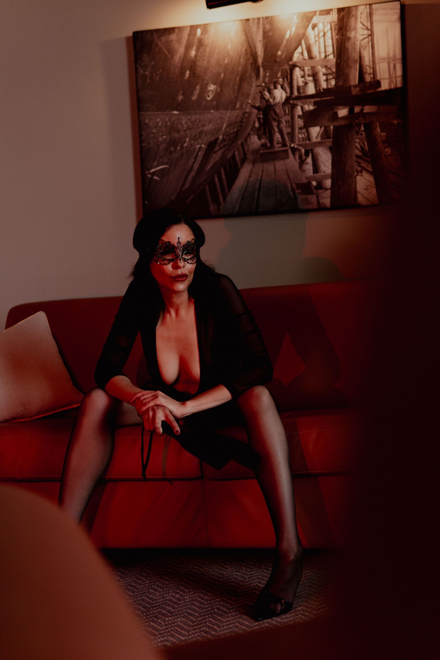Woman in black outfit and masquerade mask seated on red sofa in dimly lit room with artwork on wall.