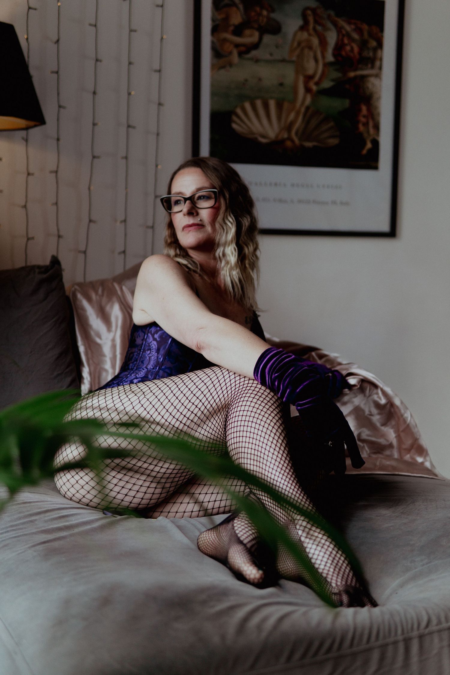 Blonde woman in purple corset with fishnet stockings and boots sitting on couch near Botticelli print. Liverpool boudoir photography