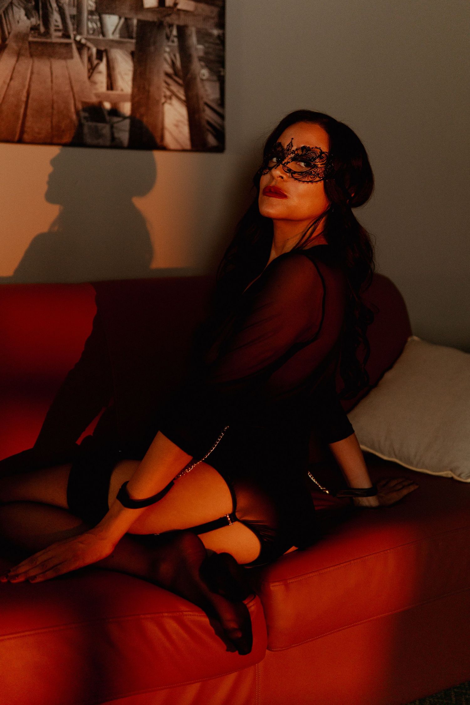 Woman in black lace mask and sheer top poses on red sofa in moody, dimly lit room.