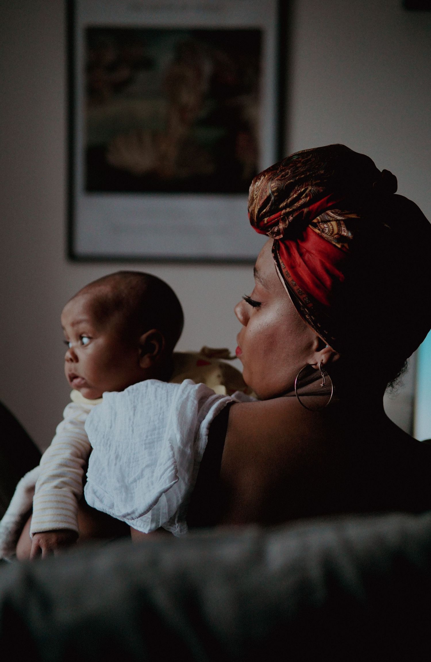 A Black mother wearing a colorful head wrap holds her infant baby while sitting indoors near a framed wall poster.
