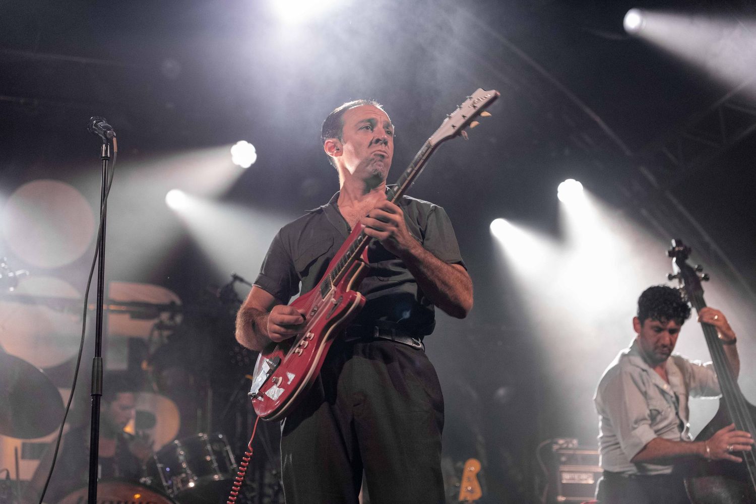 Man playing red electric guitar on stage under dramatic white spotlights during a live concert performance.