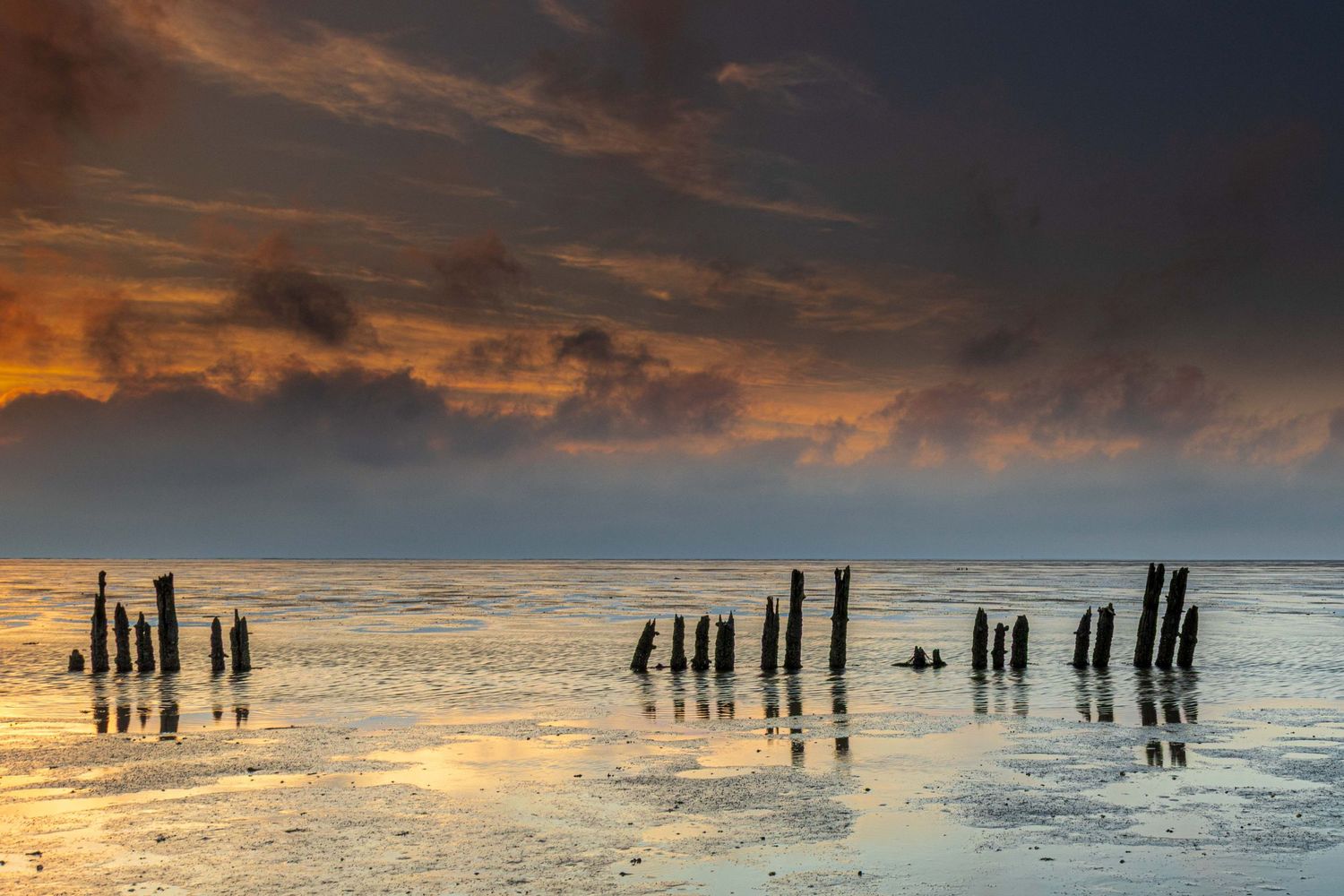 Dramatic sunset over a tidal flat with weathered wooden posts reflected in shallow water under a stormy sky.