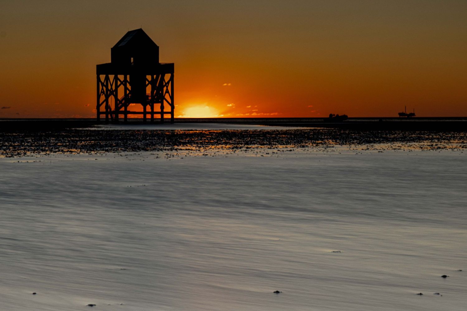 Silhouette of a coastal structure at golden sunset over calm tidal flats with a ship visible on the horizon.