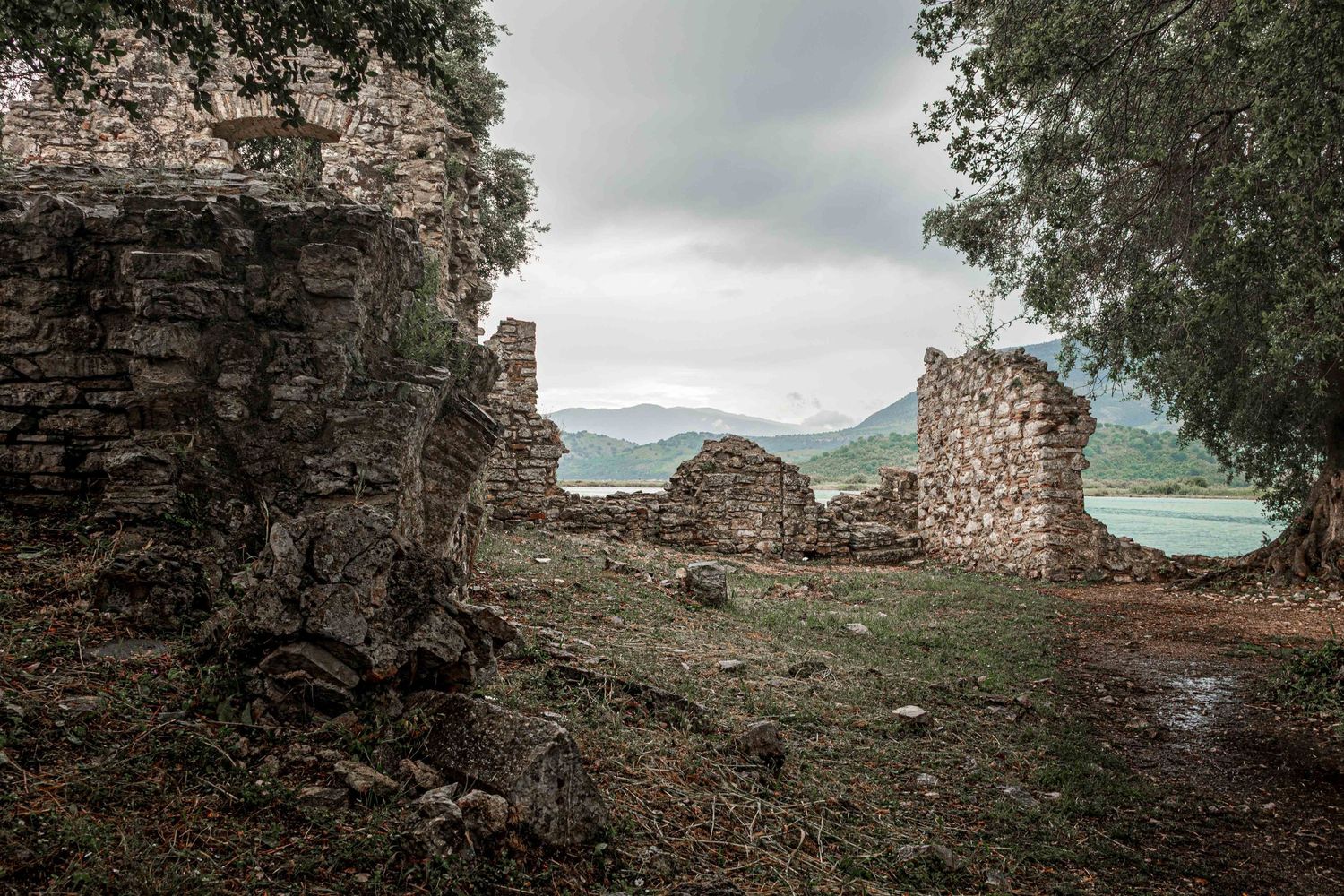 Ancient stone ruins with crumbling walls overlooking a serene lake surrounded by lush green trees under an overcast sky.