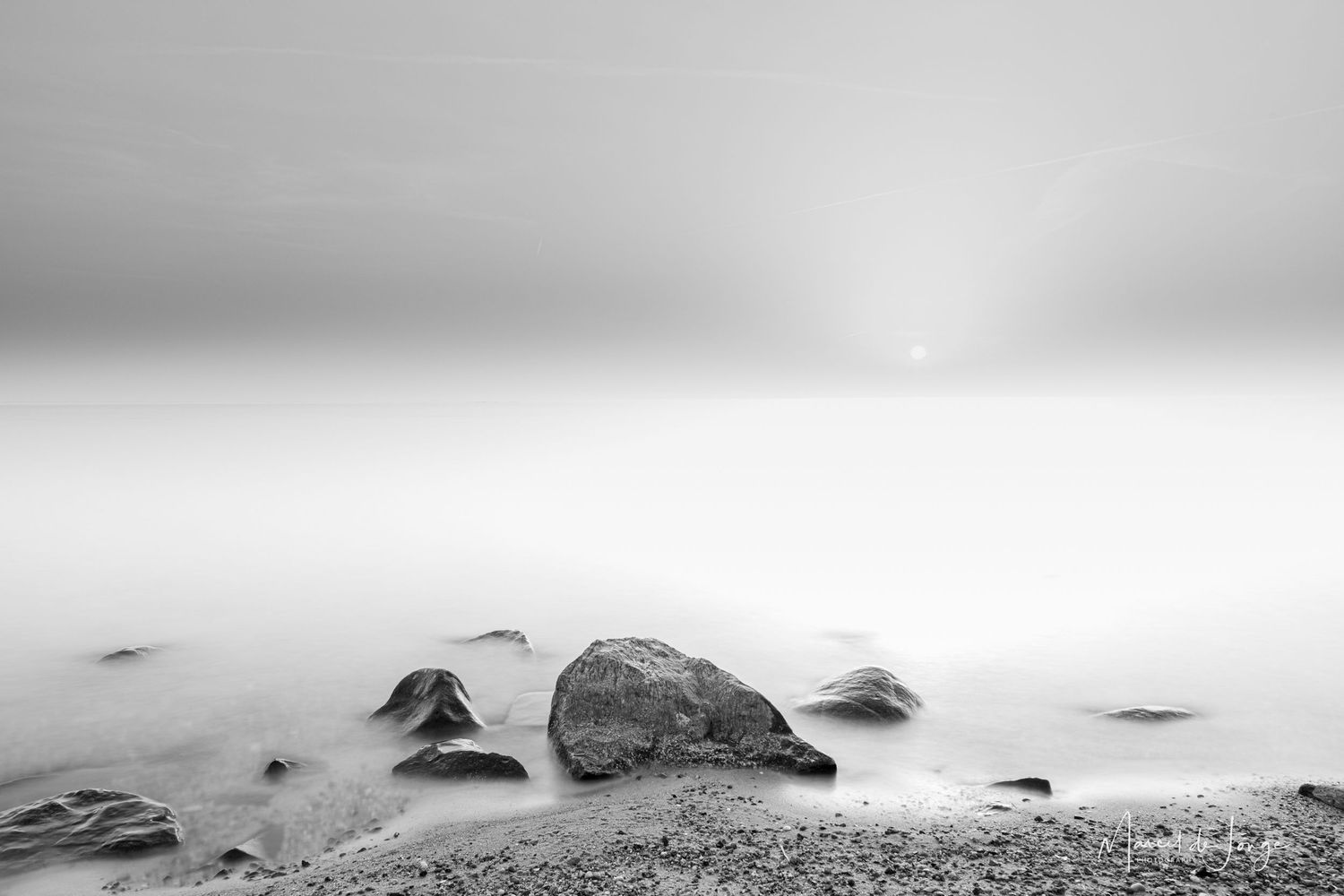 A dramatic black and white seascape shows waves crashing against rocks along a misty coastline.